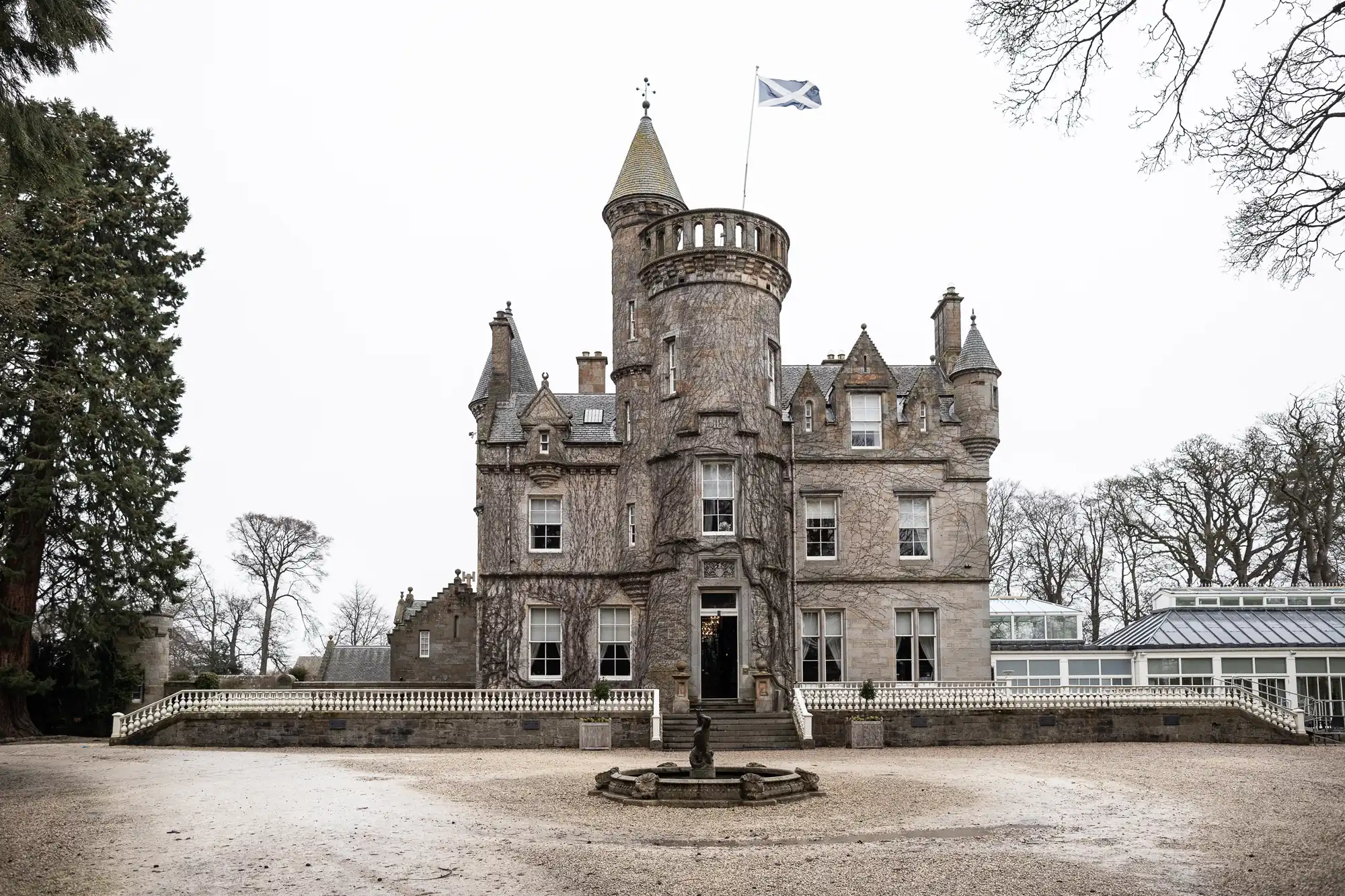 A historic stone castle with multiple turrets and a flag flying atop the central tower. The building is surrounded by bare trees and a fenced courtyard with a small fountain.
