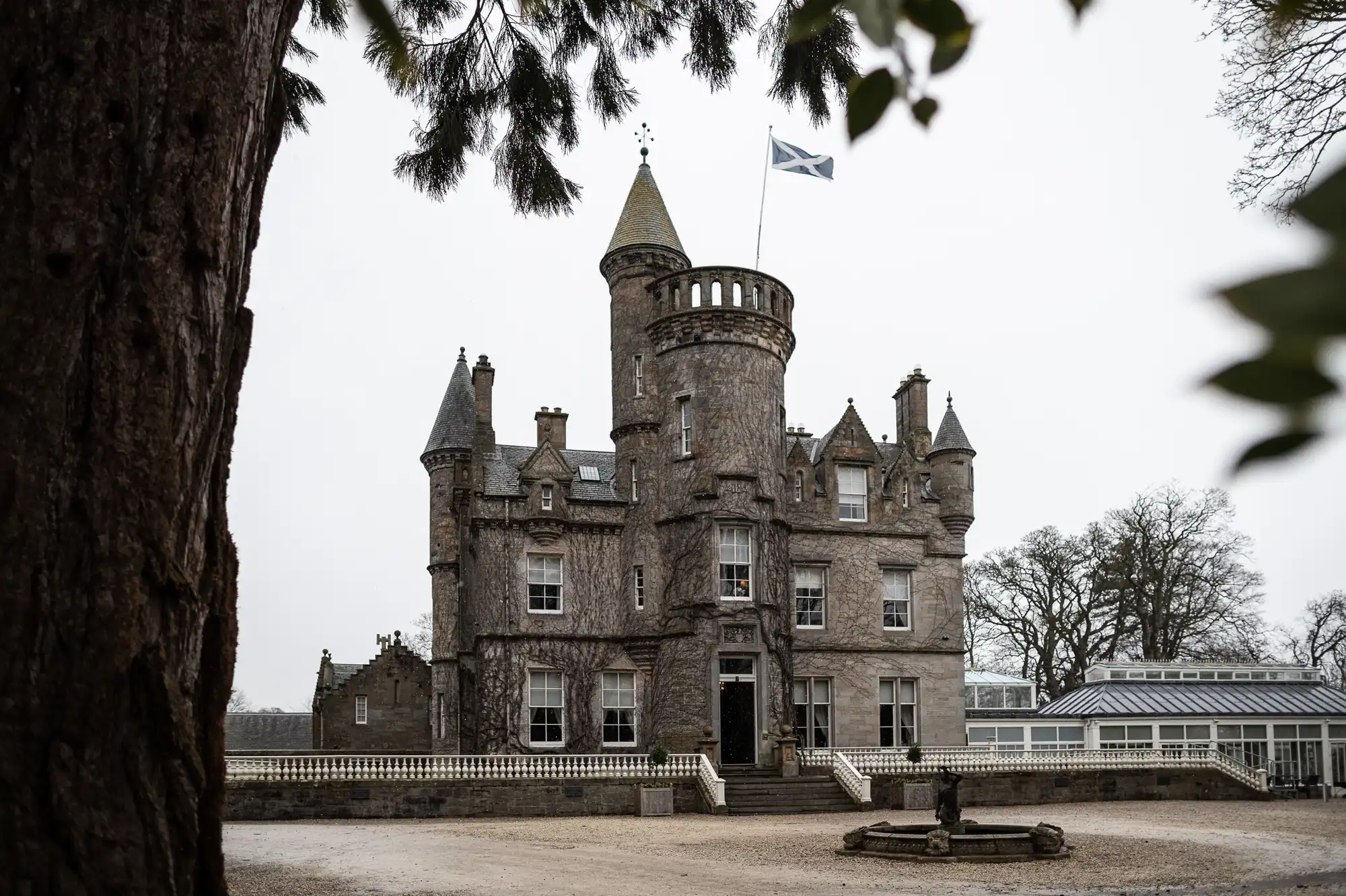 A stone castle with turrets and an entrance staircase, flying a Scottish flag, is surrounded by bare trees on a cloudy day.