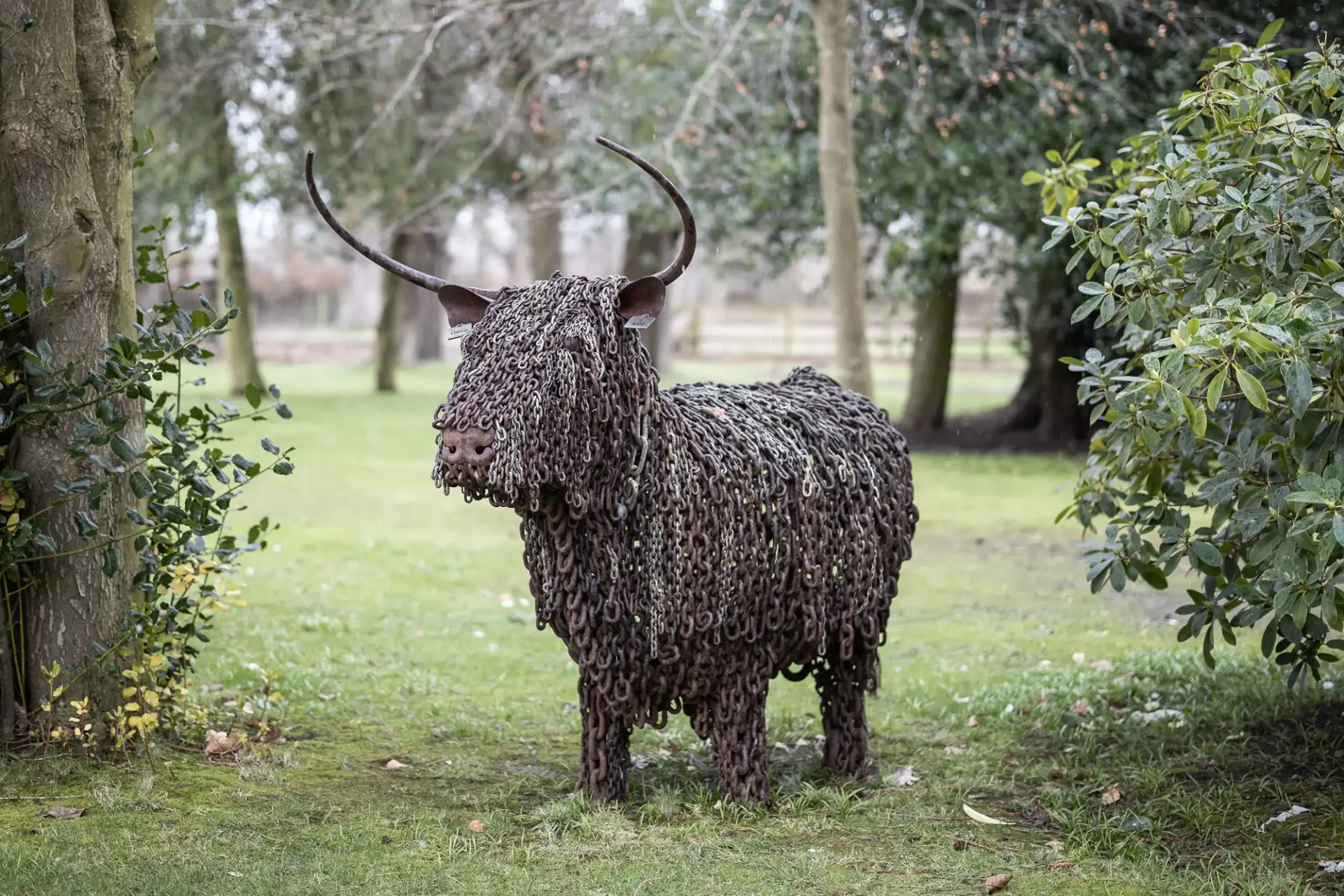 A sculpture of a bull made entirely from metal chains, displayed outdoors among trees and bushes.