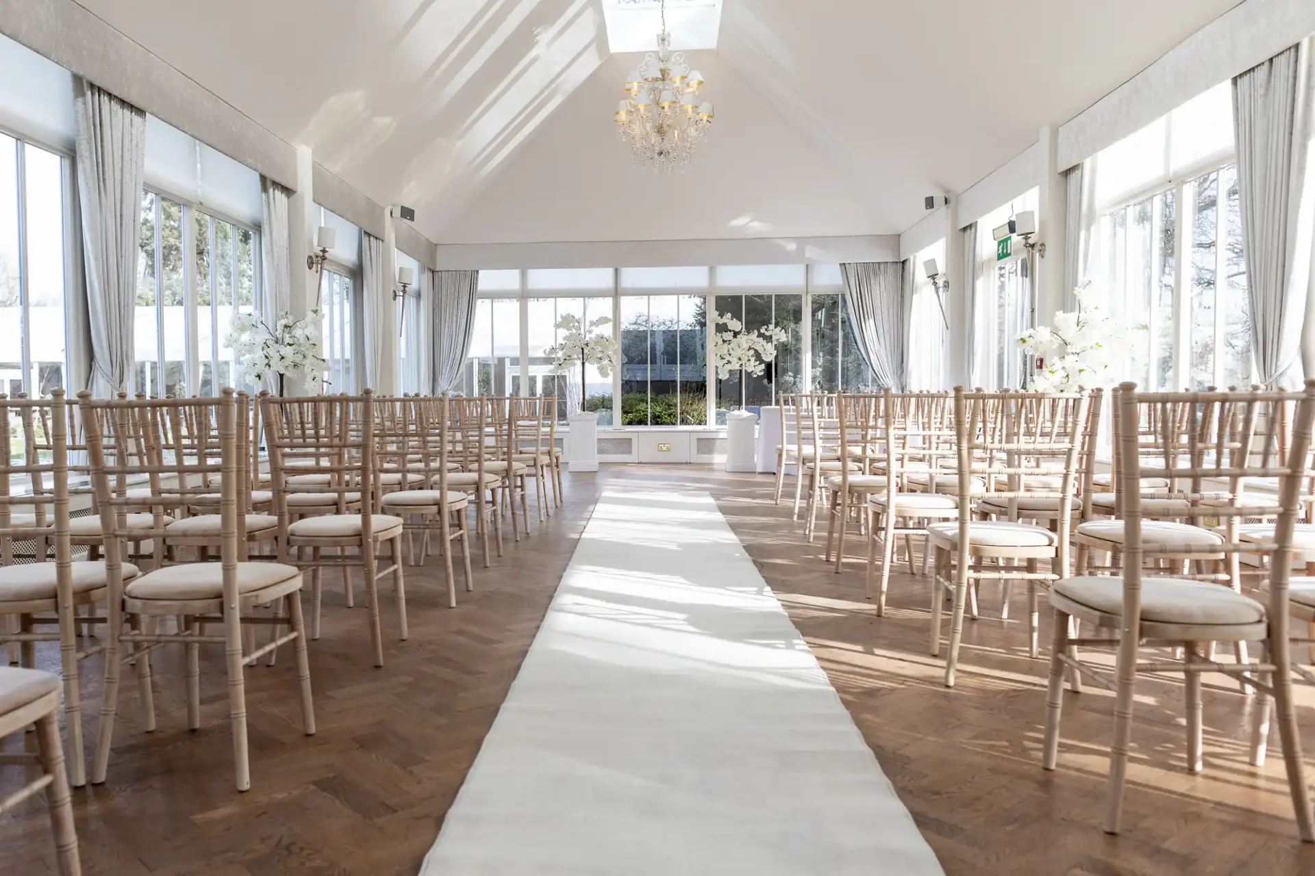 An elegant wedding ceremony room with rows of wooden chairs, a white aisle runner, and floral decorations under a chandelier, surrounded by large windows.