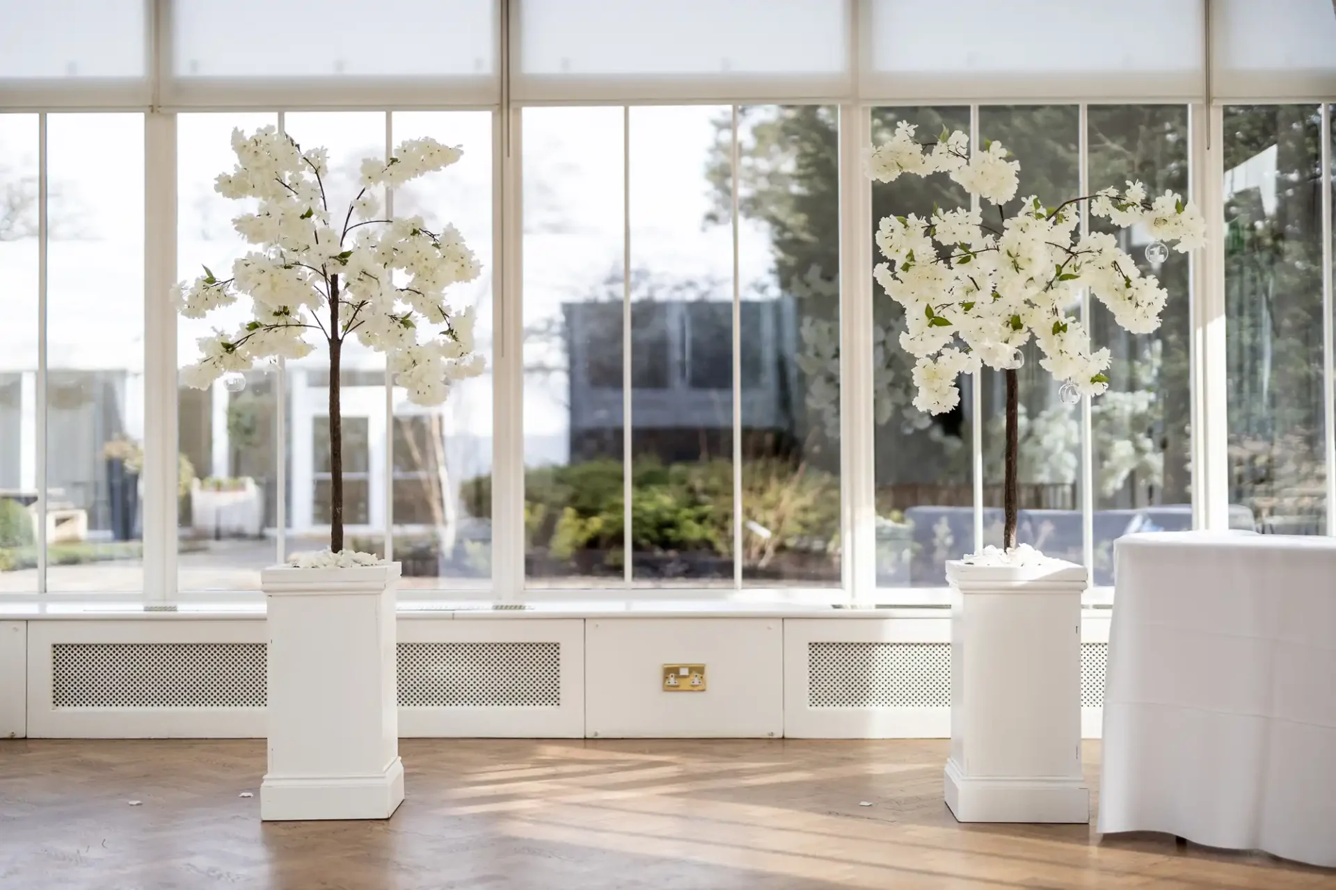 Two white floral arrangements on pedestals in a bright room with large windows overlooking a garden.