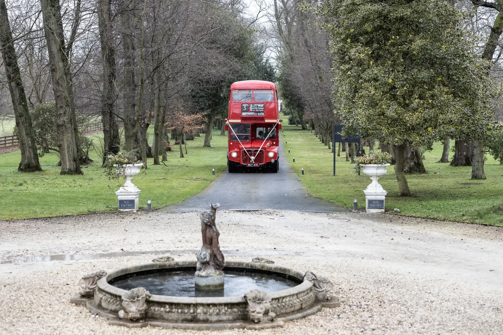 A red double-decker bus drives along a tree-lined gravel path with a small stone fountain in the foreground.