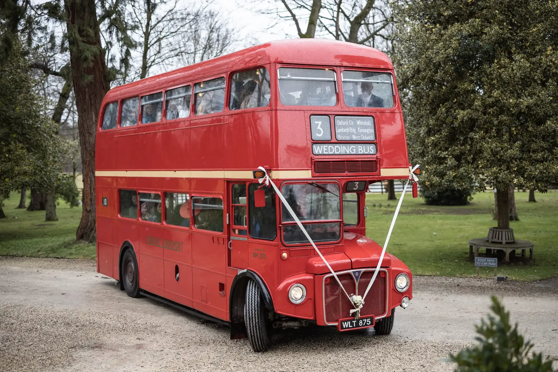 A red double-decker bus labeled "wedding bus" parked on a gravel path in a serene park setting with trees in the background.
