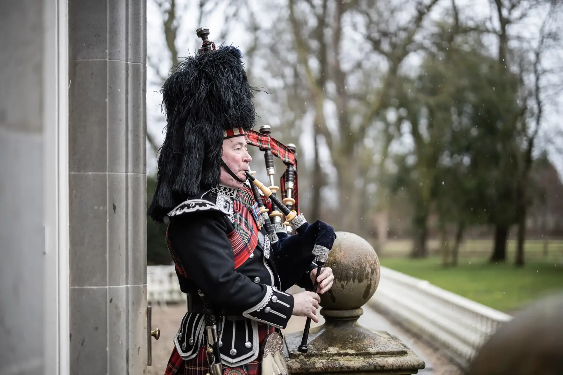 A bagpiper in traditional scottish attire playing outdoors near a stone building, wearing a black feathered hat and kilt.