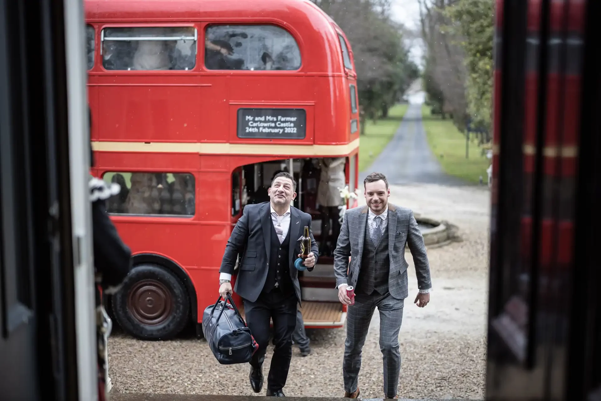 Two men in suits joyfully running with champagne and a bag toward a red double-decker bus parked on a gravel path, viewed from inside another vehicle.