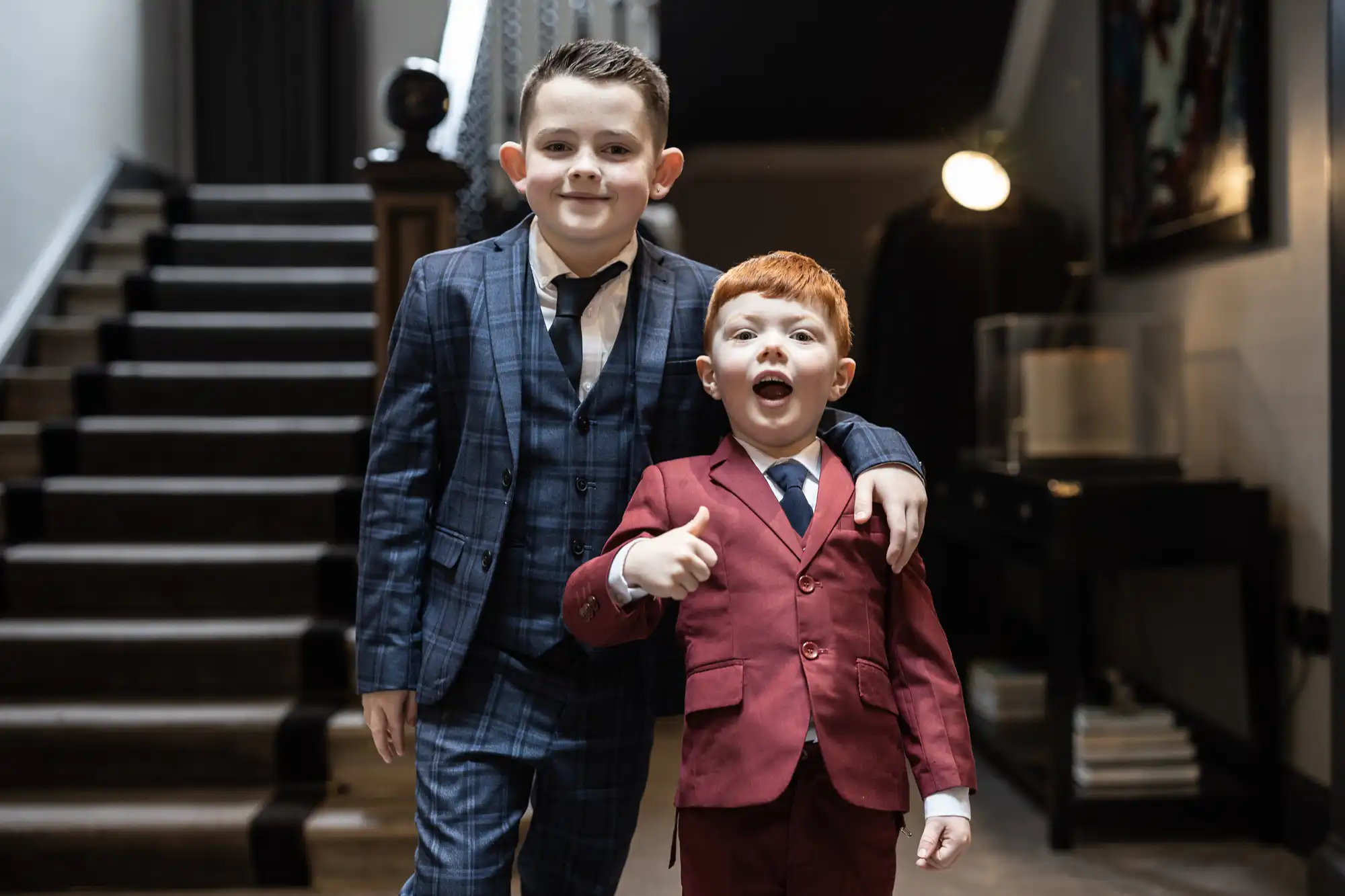 Two young boys dressed in suits stand inside a modern home, with one boy in a blue plaid suit and the other in a red suit giving a thumbs up gesture.