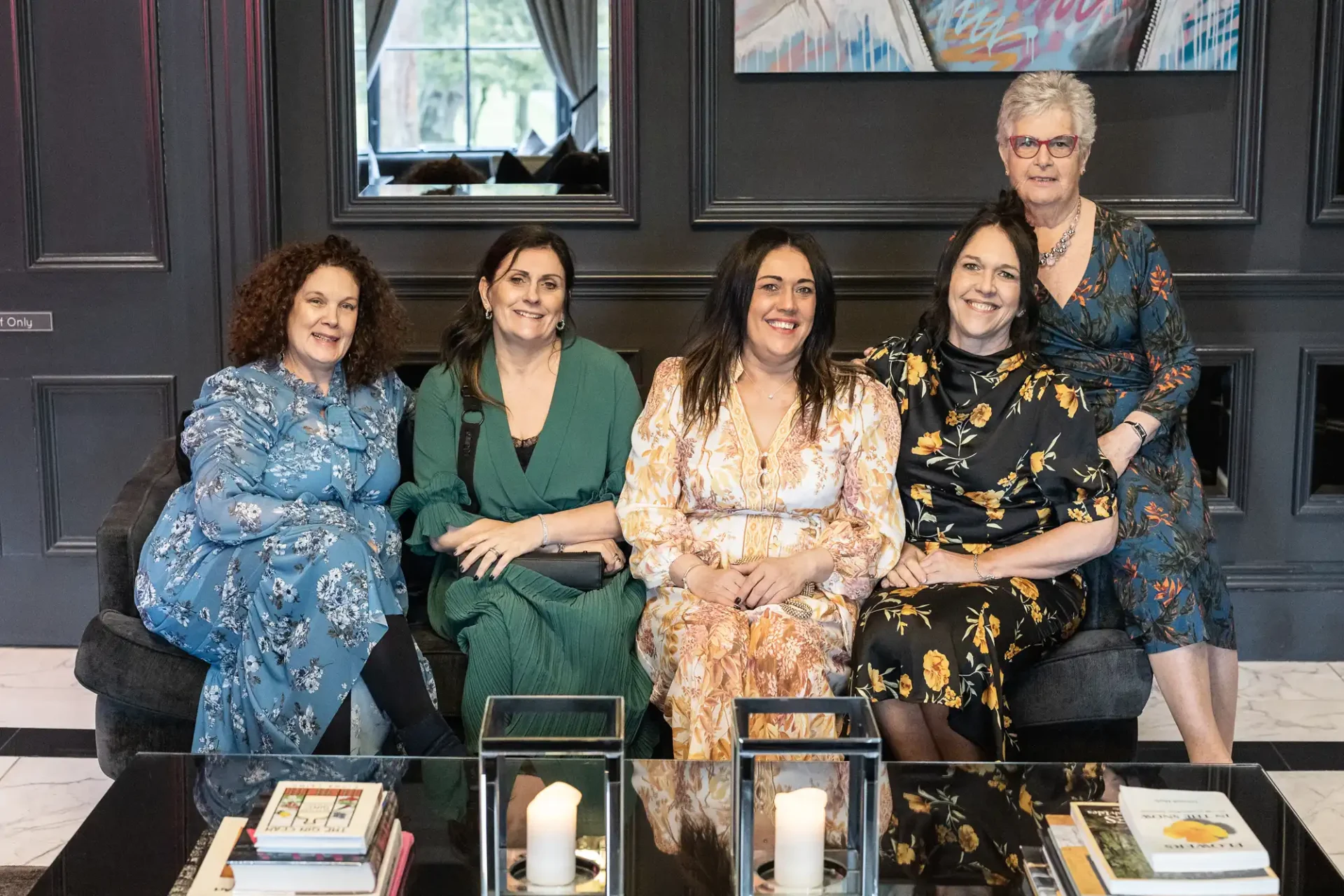 Five women smiling, seated on a sofa in a stylish room with artworks and a coffee table decorated with books and candles.