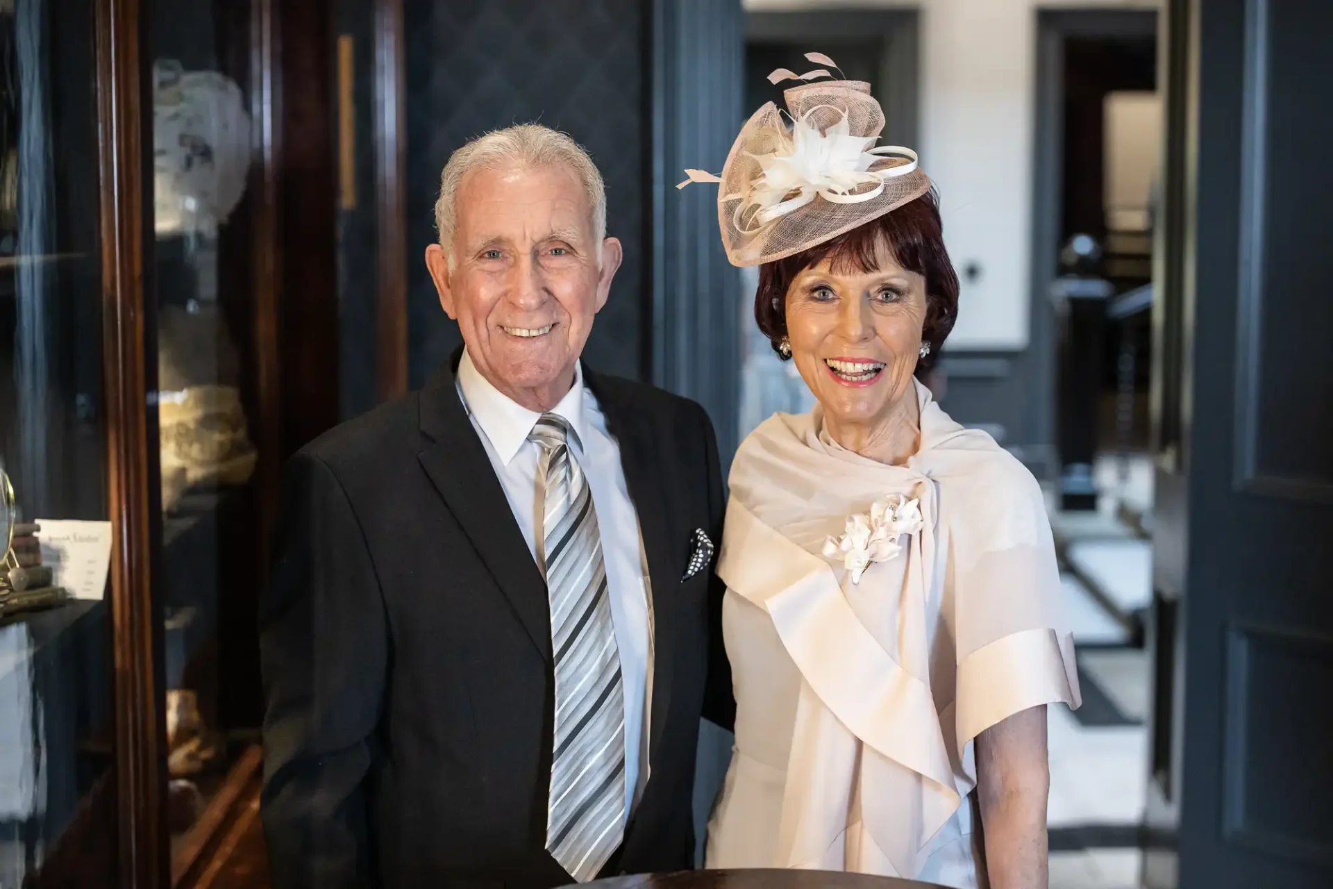 An elderly couple dressed in formal attire smiling indoors, the woman wears a hat with a feather decoration, and the man is in a suit and tie.