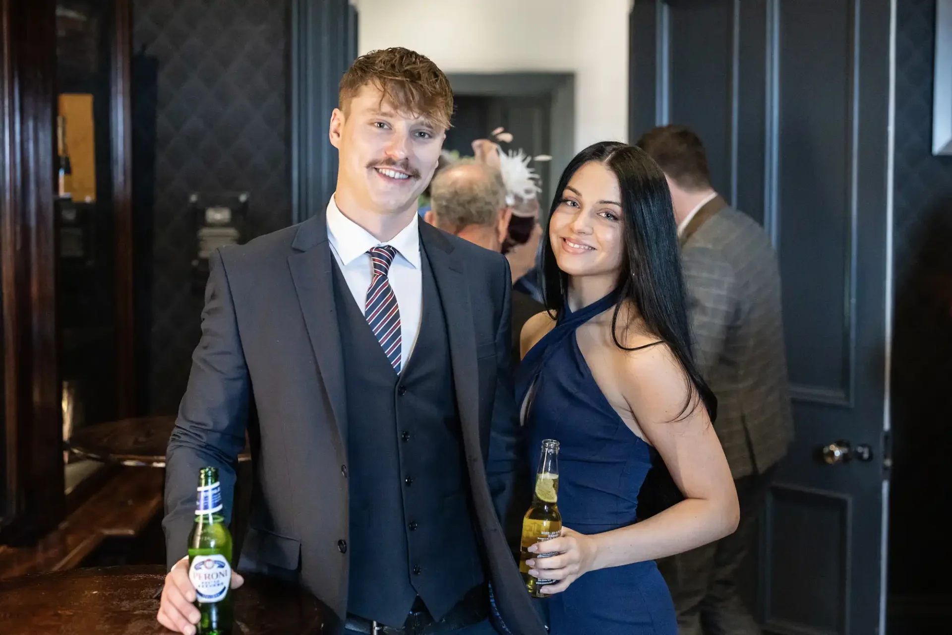 A young couple smiling at the camera, each holding a bottle of beer, at a formal indoor event.