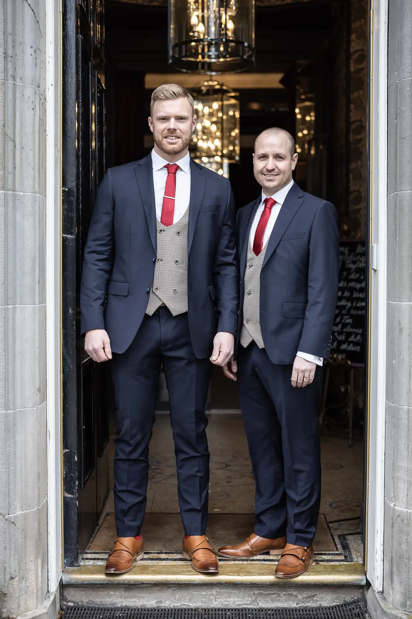 Two men in business suits and red ties standing in a doorway, smiling at the camera.