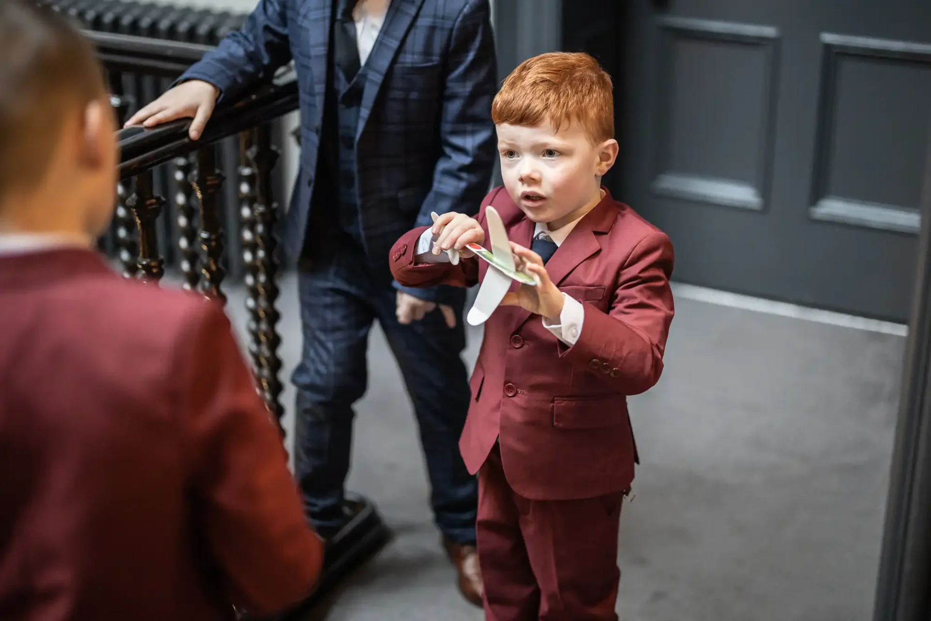 A young boy in a burgundy suit holding a paper airplane indoors, talking to another boy, with an adult in the background.