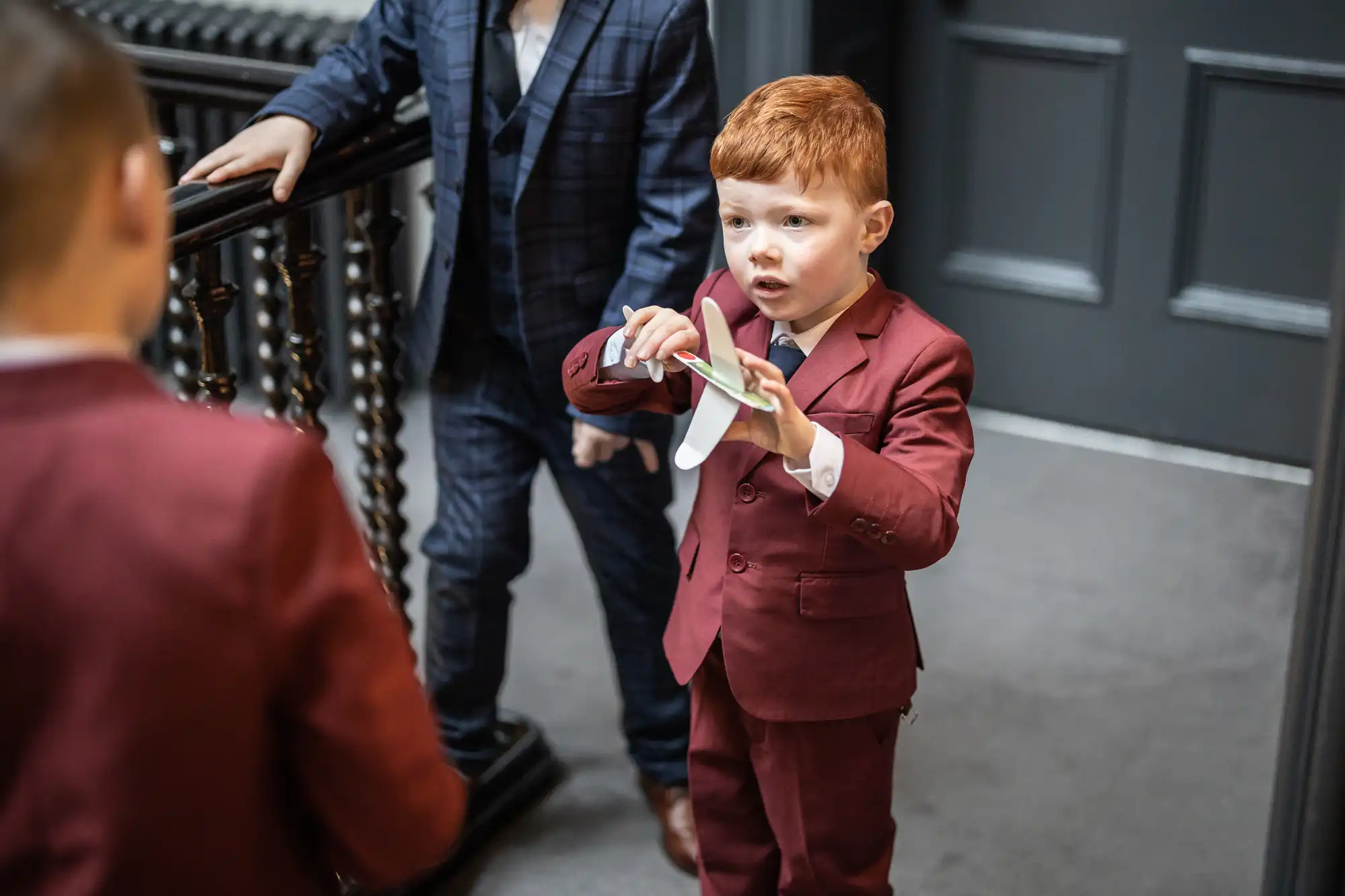 A young boy in a maroon suit holds a toy while talking to another child in a similar suit. A third child, dressed in a navy suit, stands next to a staircase railing in the background.