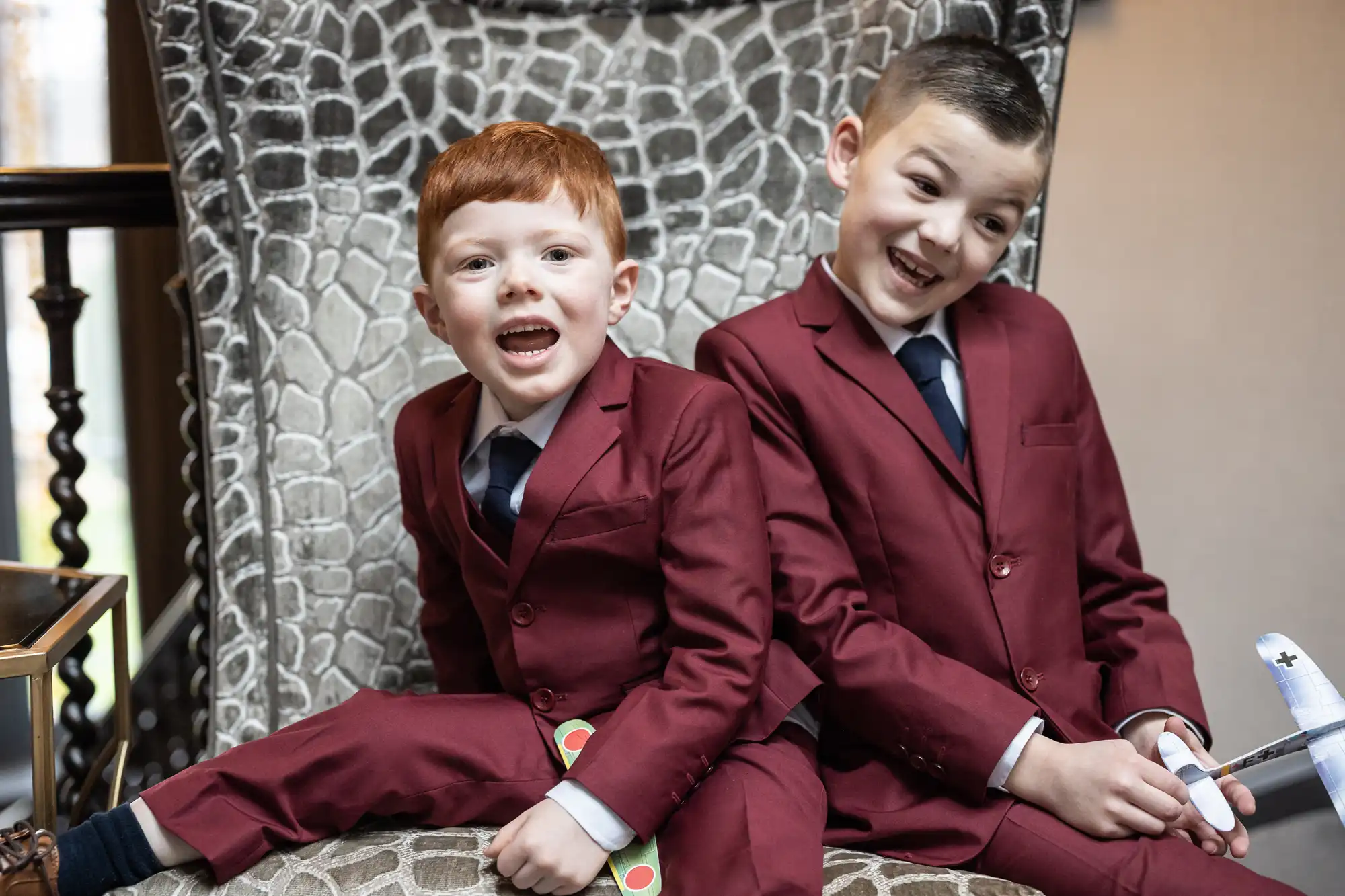 Two boys in matching maroon suits and blue ties sit on a patterned chair, smiling and holding toys.