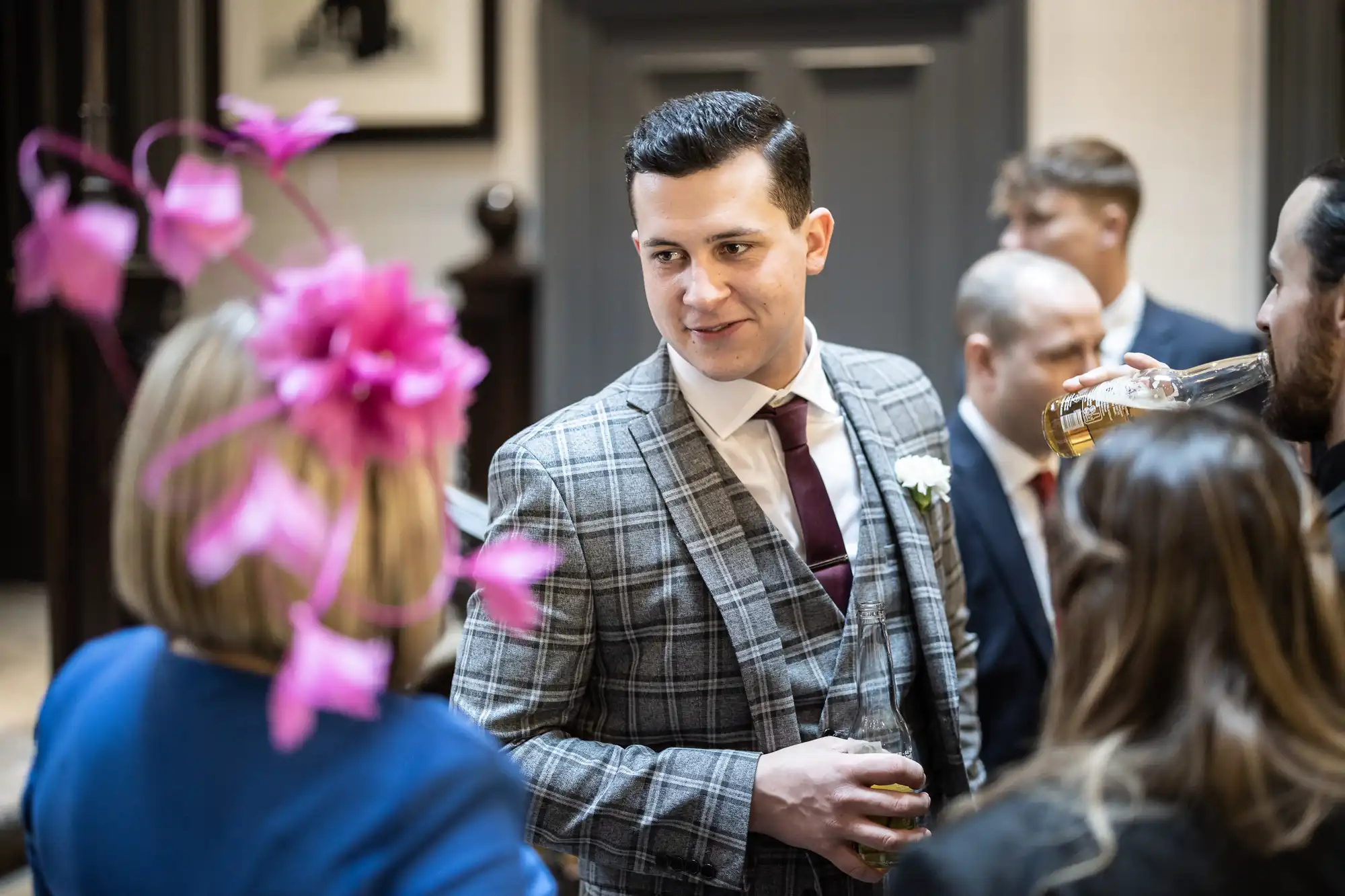 A man in a grey plaid suit and maroon tie holds a drink, conversing with a female guest in a blue dress and pink floral headpiece at a social event. Other guests can be seen in the background.