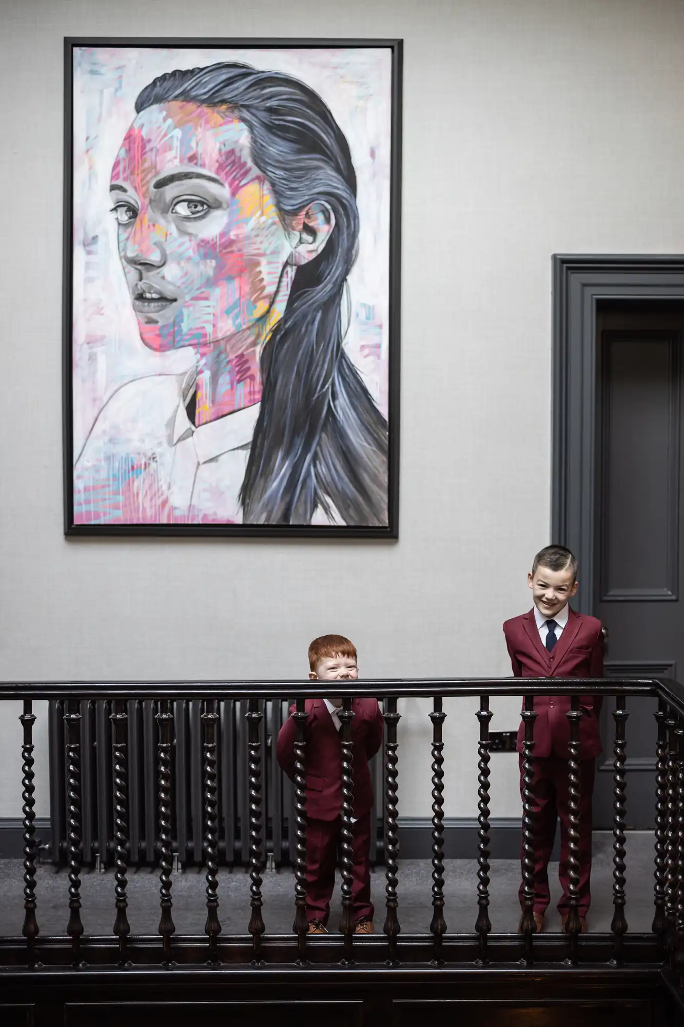 Portrait of a woman with vibrant pink and blue streaks on canvas, displayed above a staircase with two young boys in burgundy suits observing it.