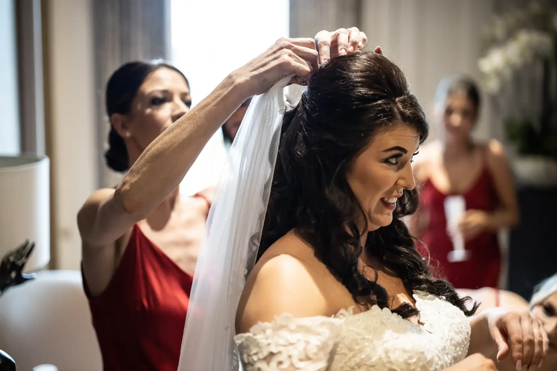 A bride smiles as another woman adjusts her veil in a room, with a bridesmaid in a red dress in the background.