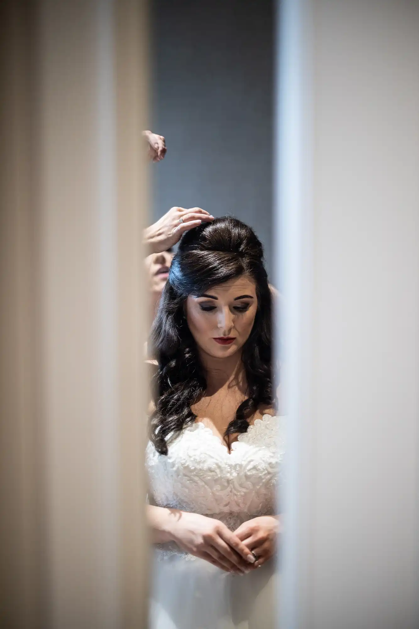 A bride in a white dress is reflected in a mirror as she adjusts her hair, looking downwards thoughtfully.
