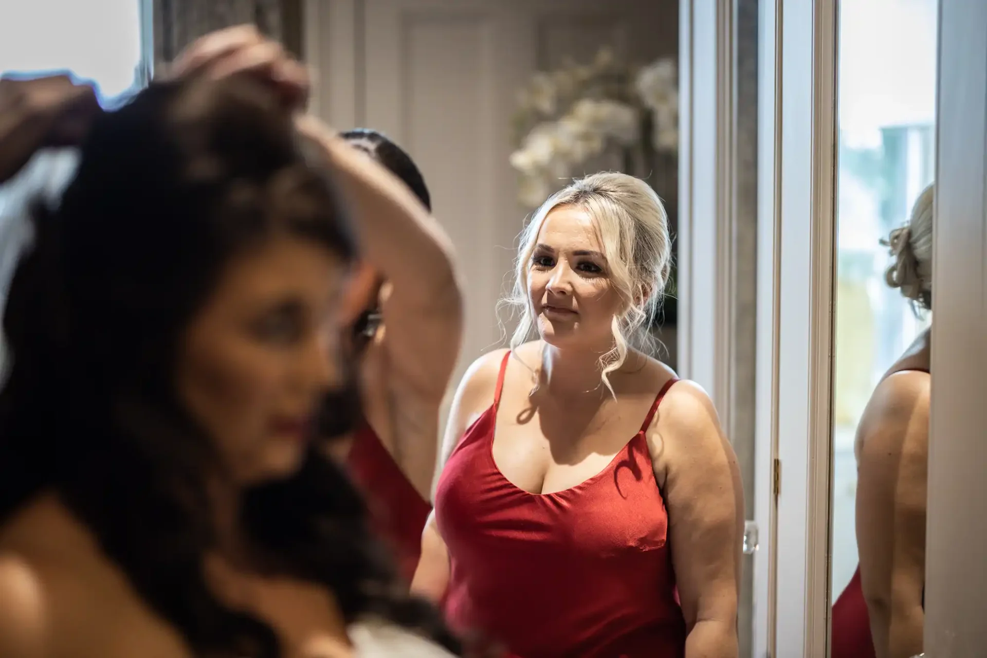 A woman in a red dress looks fondly at another woman adjusting her hair in a mirror-lit room.