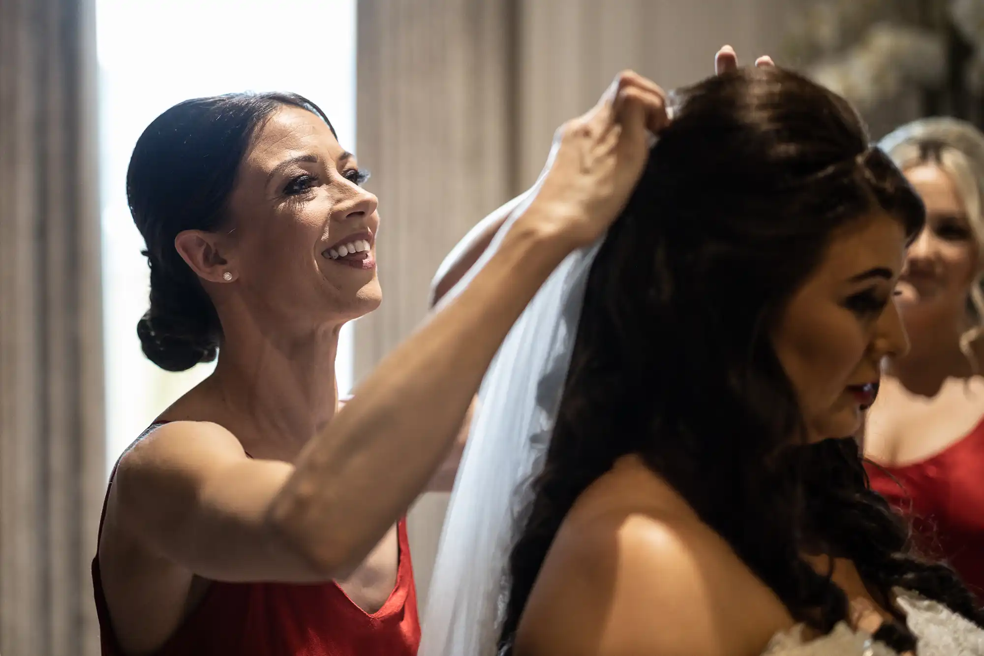 A woman in a red dress helps another woman adjust her wedding veil while another woman looks on.