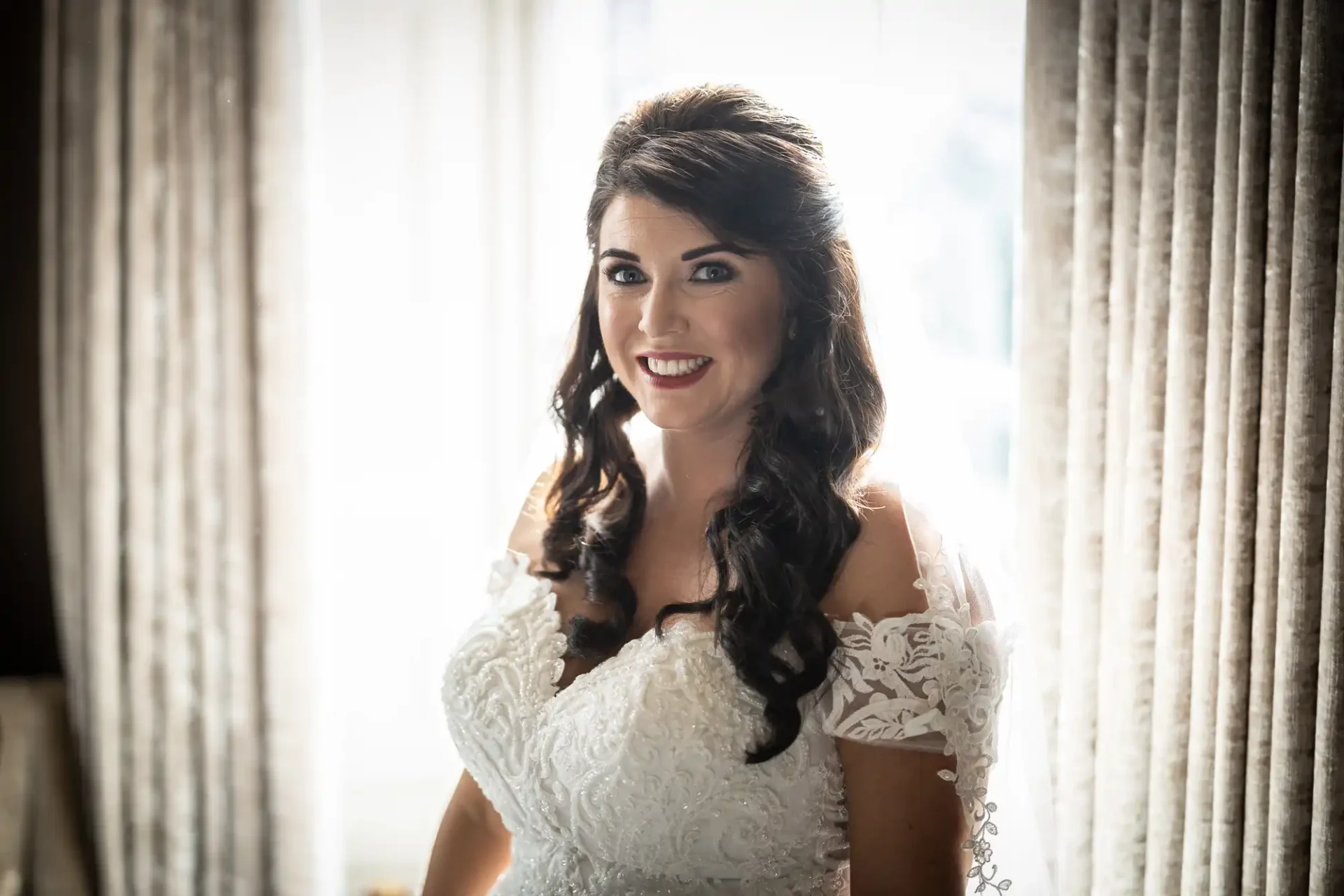 A bride in a white lace dress smiles warmly, standing near a window with sheer curtains diffusing light behind her.