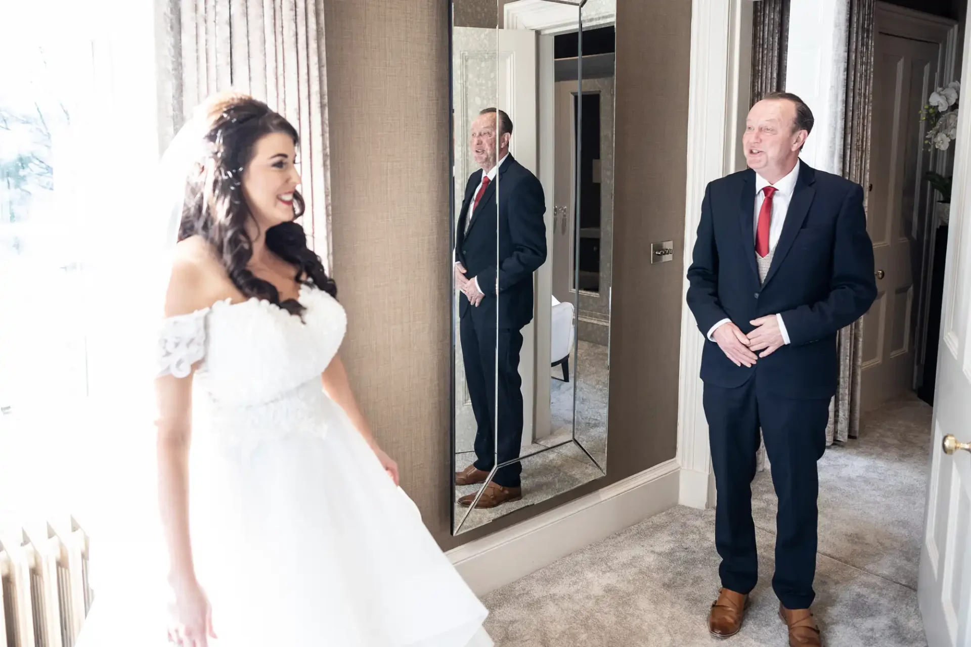 A bride in a white dress smiles at her father, who is reflected in a mirror, in a well-lit room.