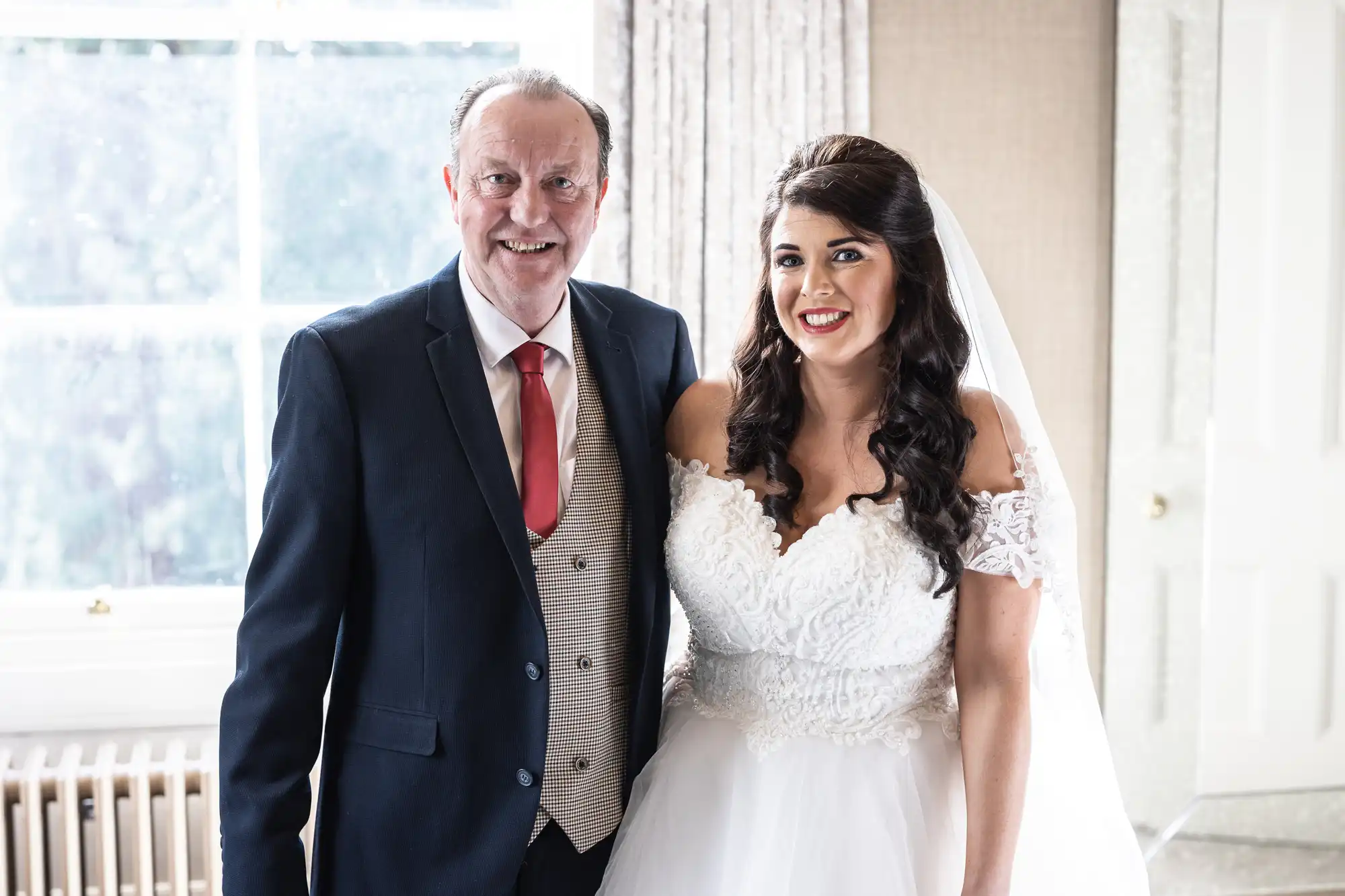 A man in a suit and tie stands beside a woman in a white wedding dress, both smiling, in a brightly lit room with a window in the background.
