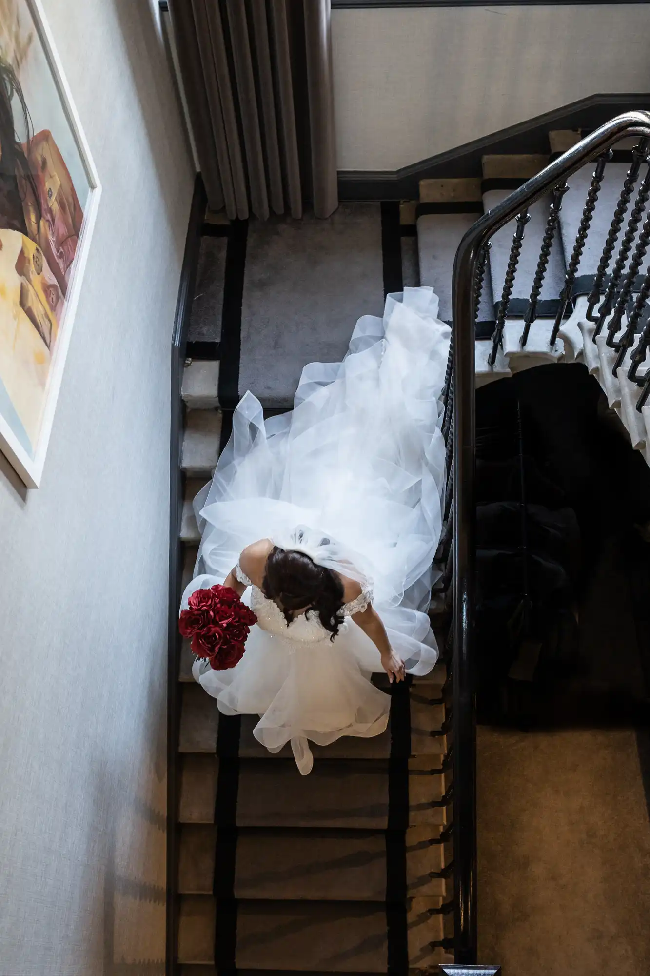 Bride in a white dress with a long train holding a red bouquet ascends a spiral staircase, viewed from above.