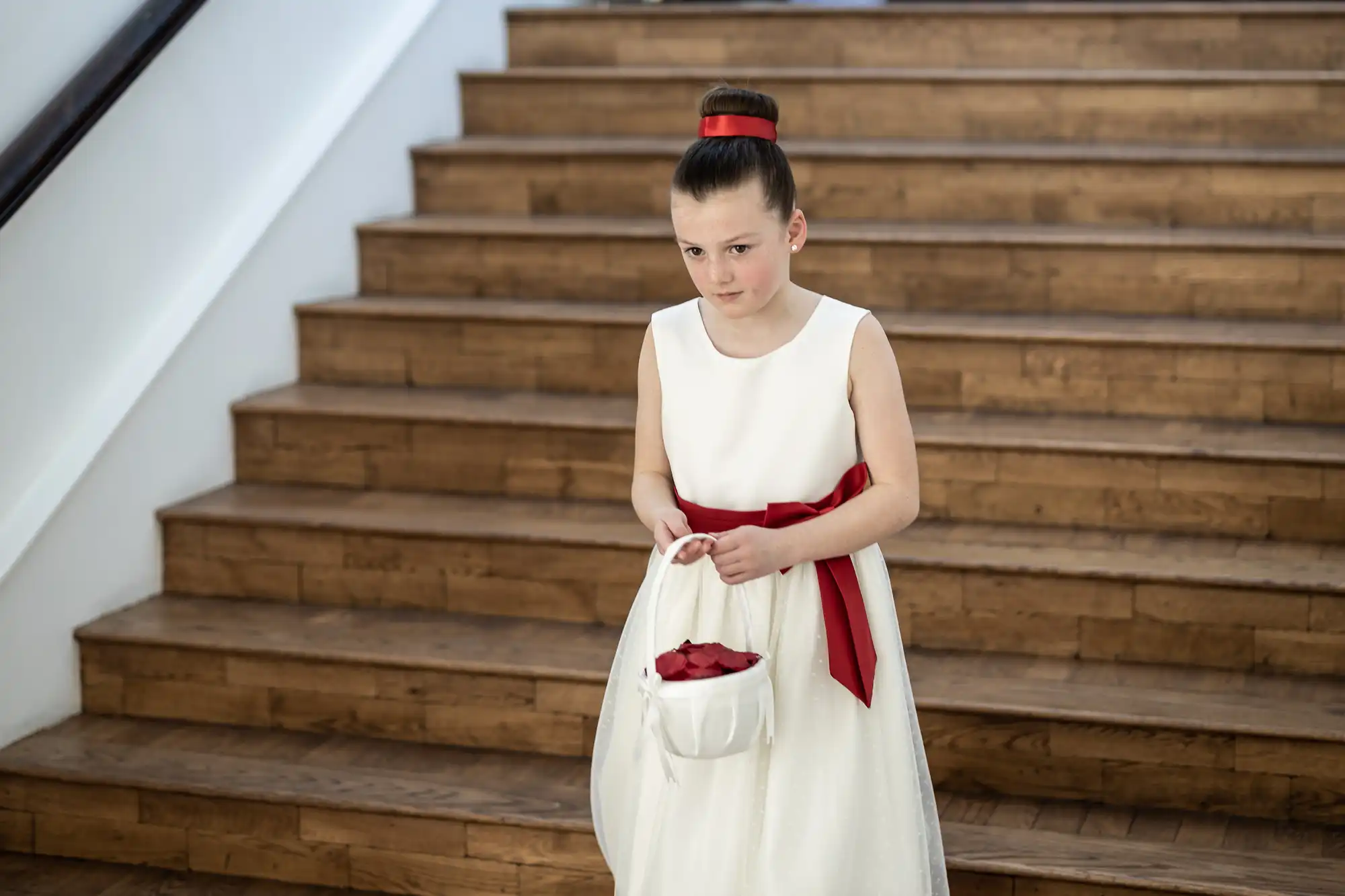 A young girl in a white dress with a red sash stands holding a white basket with red petals, posed in front of a wooden staircase.