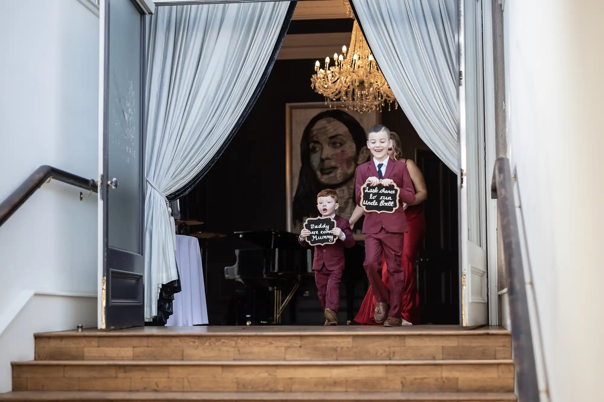 Two children dressed in matching maroon suits walk down a staircase, each holding a sign. The interior shows draped curtains and a chandelier in the background.