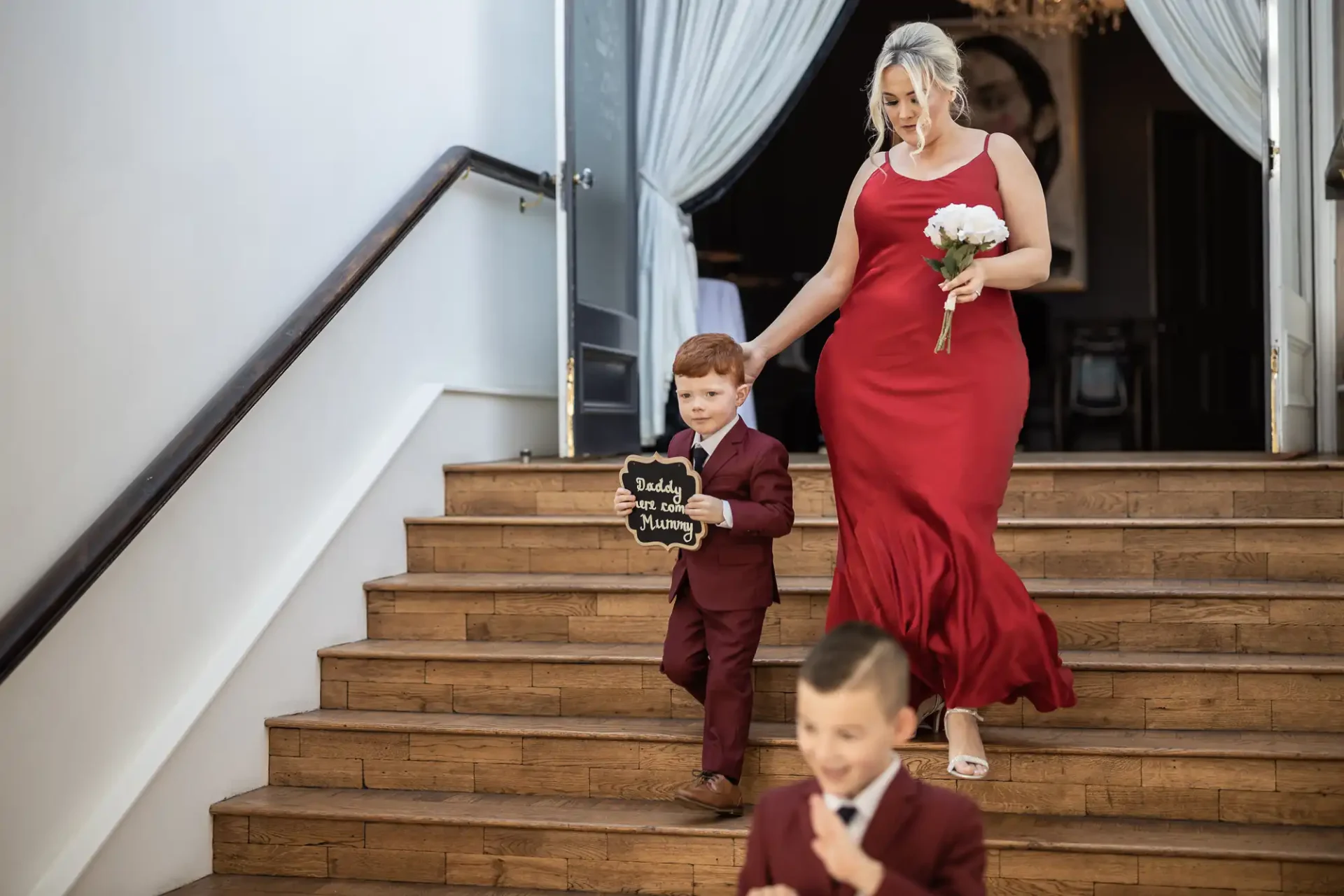 A woman in a red dress and a young boy in a suit descending stairs, with the boy holding a "daddy here comes mummy" sign. another boy runs in the foreground.