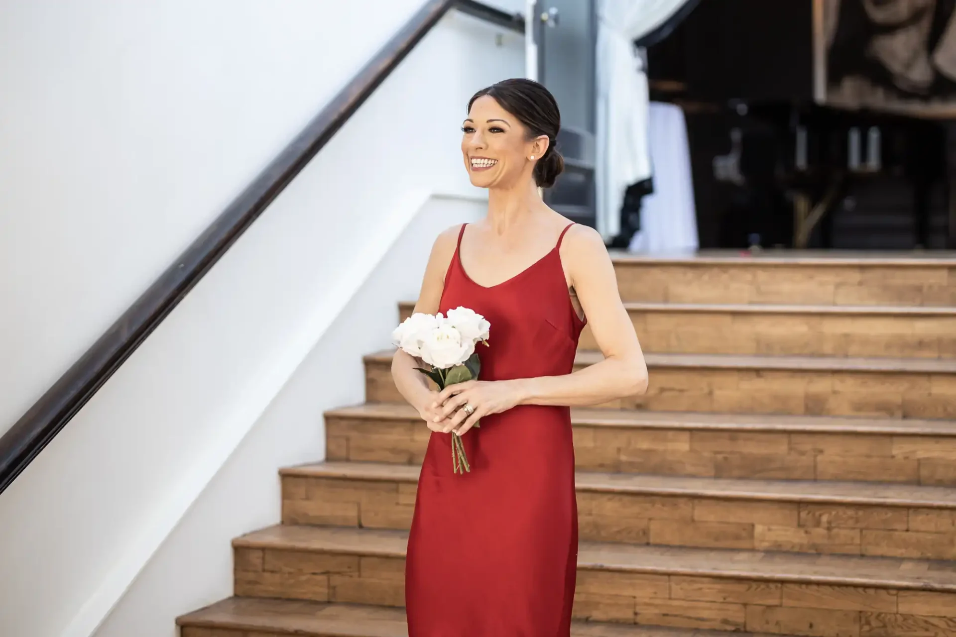 A woman in a red dress smiles while holding a bouquet of white flowers, standing on a staircase.