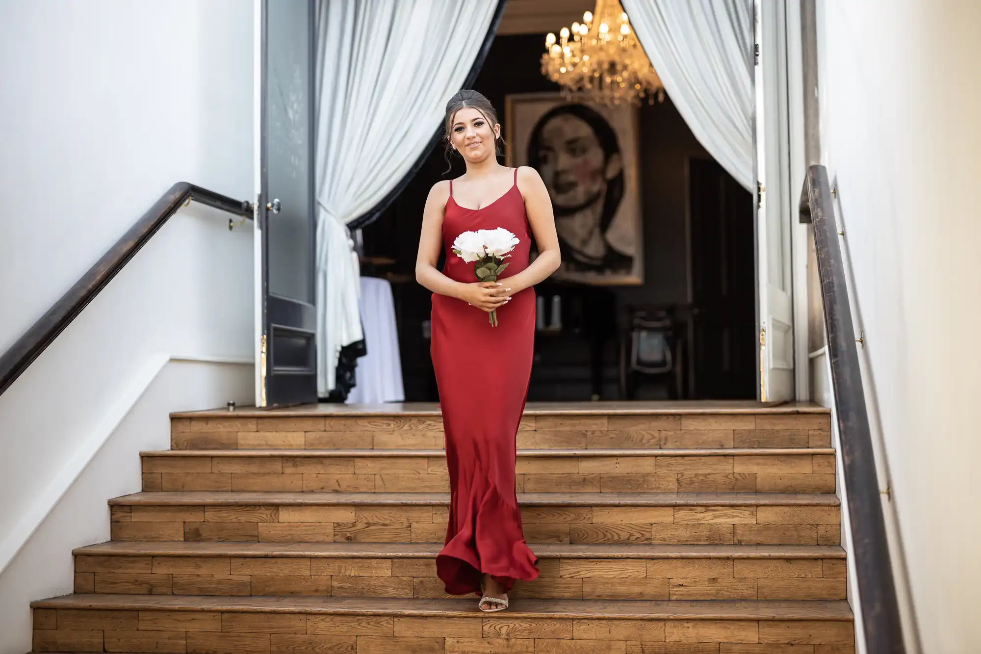 A woman in a red dress holds a bouquet of white flowers while standing on a wooden staircase with a chandelier and large artwork in the background.