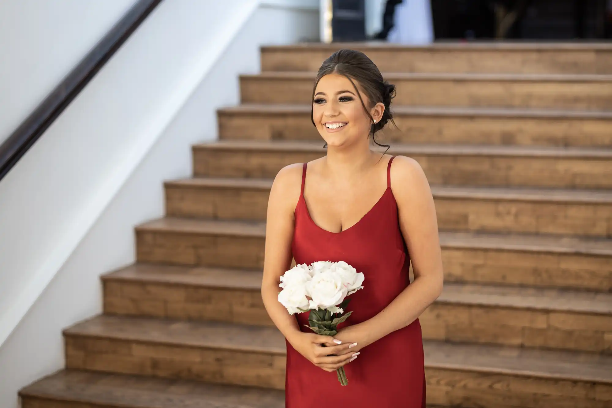 A woman in a red dress holds a bouquet of white flowers while standing on a wooden staircase, smiling.