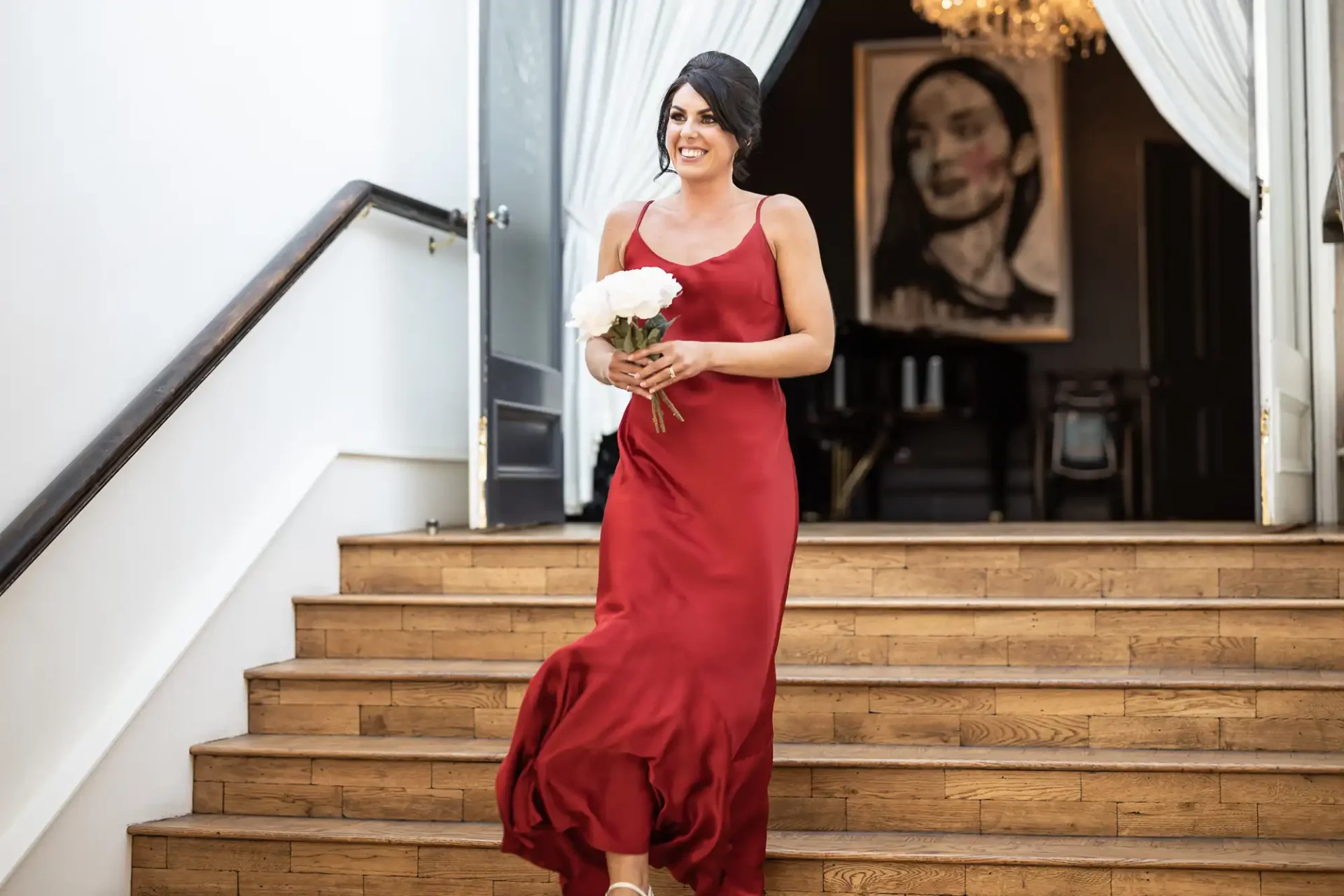 A woman in a red dress smiling as she descends a staircase, holding a bouquet, with a portrait in the background.