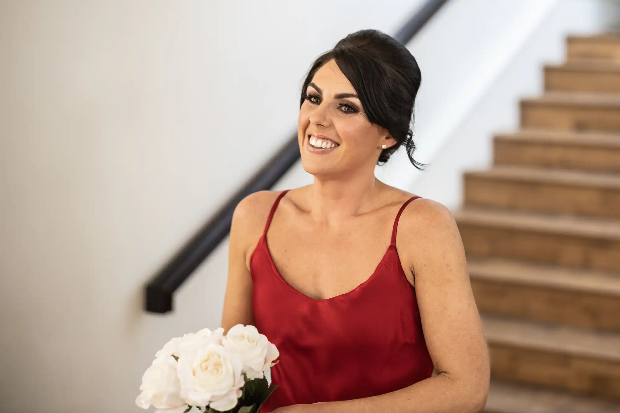 A woman in a red dress holds white flowers and smiles while standing near a staircase.