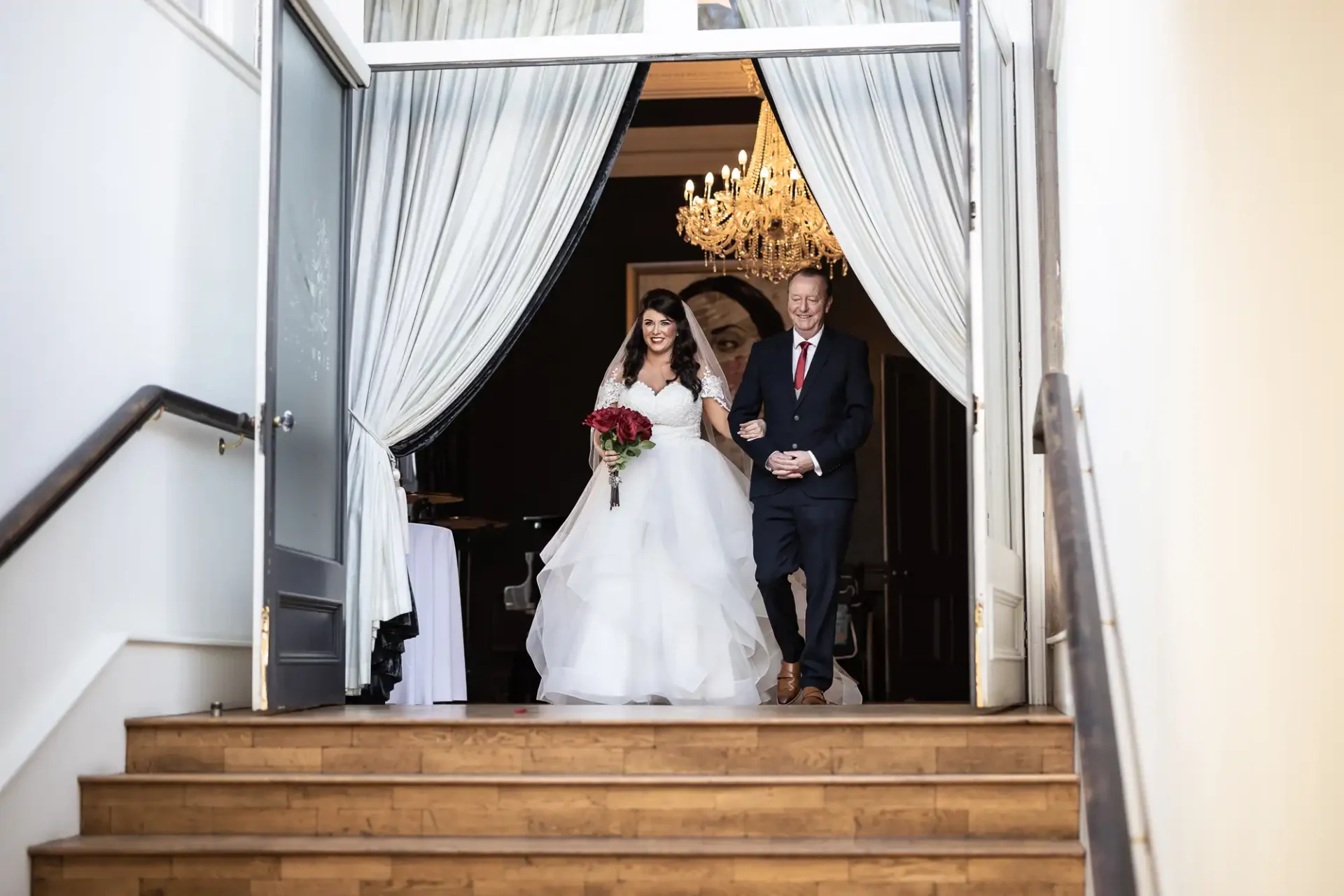 A bride and groom smiling as they descend a staircase, the bride holding a bouquet, with an elegant chandelier above.