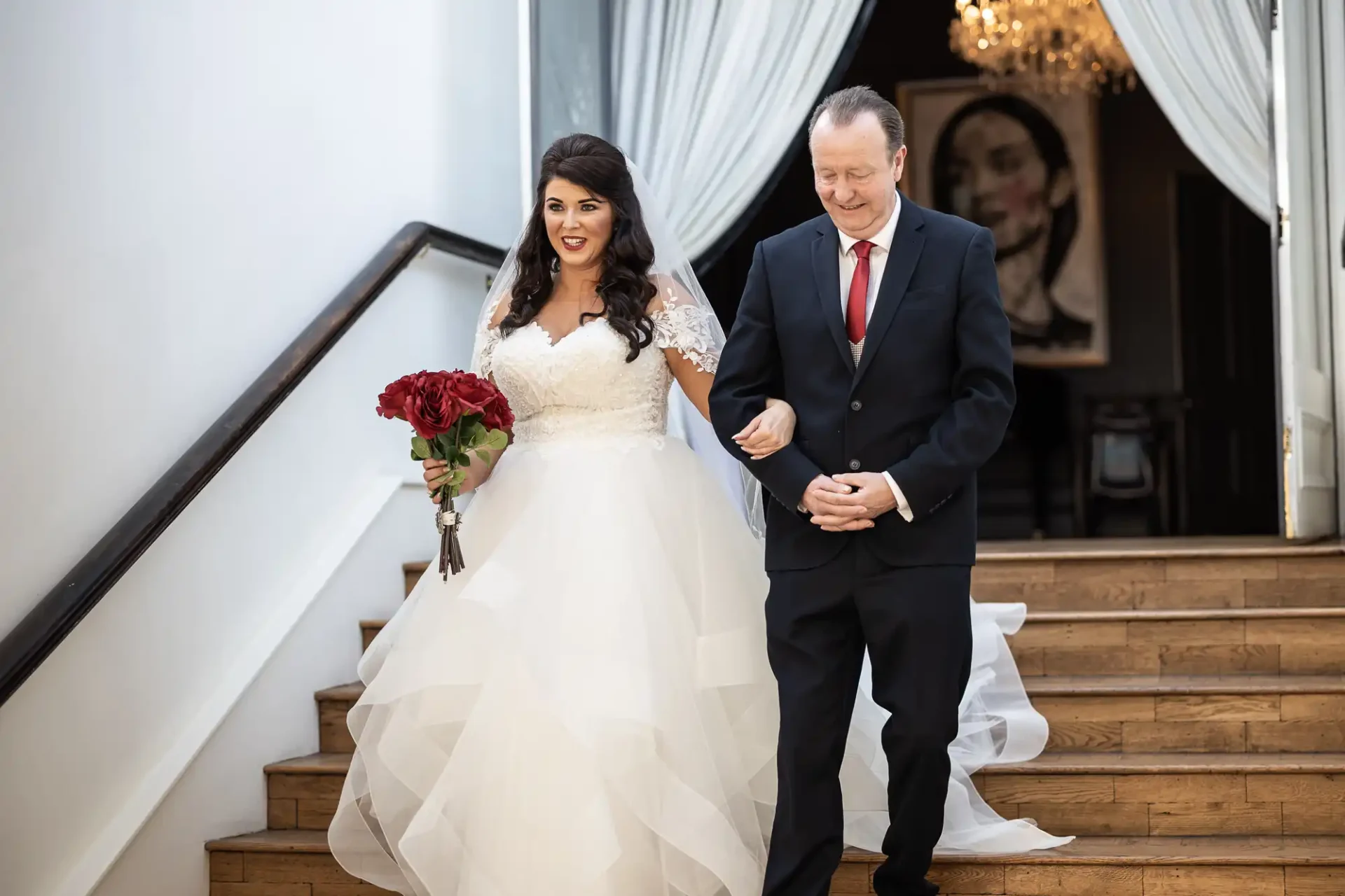 Bride and father descend a staircase, smiling, at a wedding venue. she holds a red bouquet; both are dressed formally.