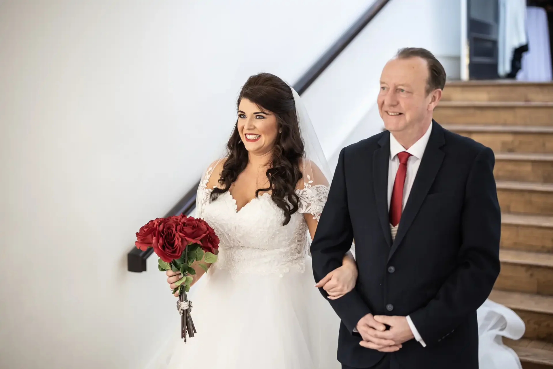 Bride in a white dress holding a bouquet of red roses walks down a staircase with an older man in a suit, both smiling.