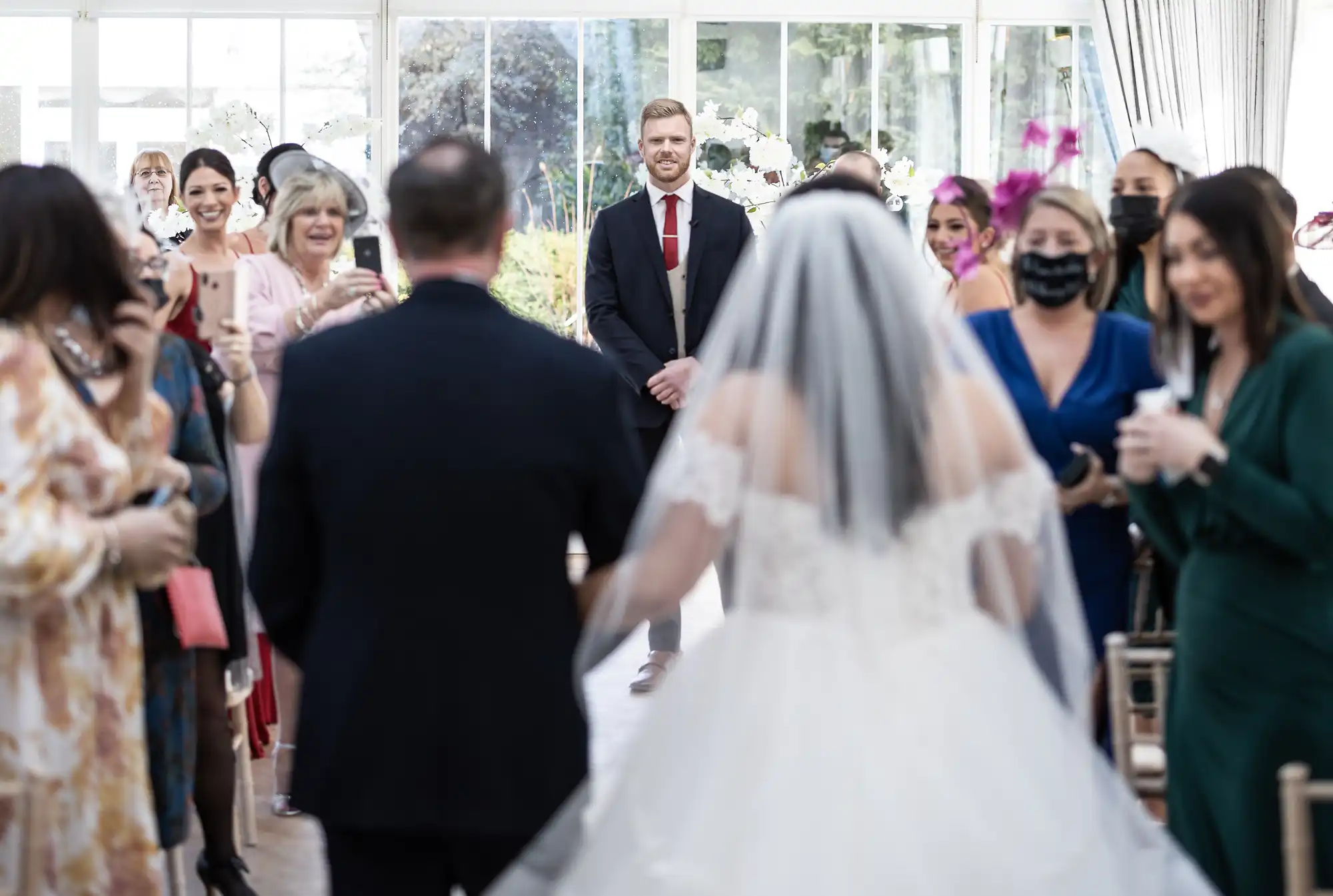 A bride walks down the aisle towards the groom, who stands at the altar. Guests, some taking photos, watch the scene inside a bright wedding venue.