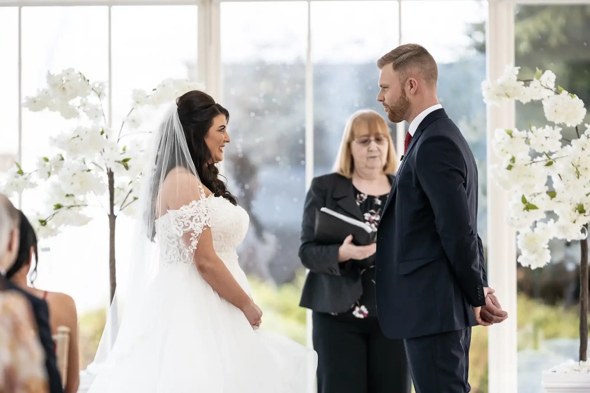 A bride and groom exchange vows in a sunlit room with white floral decorations, officiant and guests in attendance.
