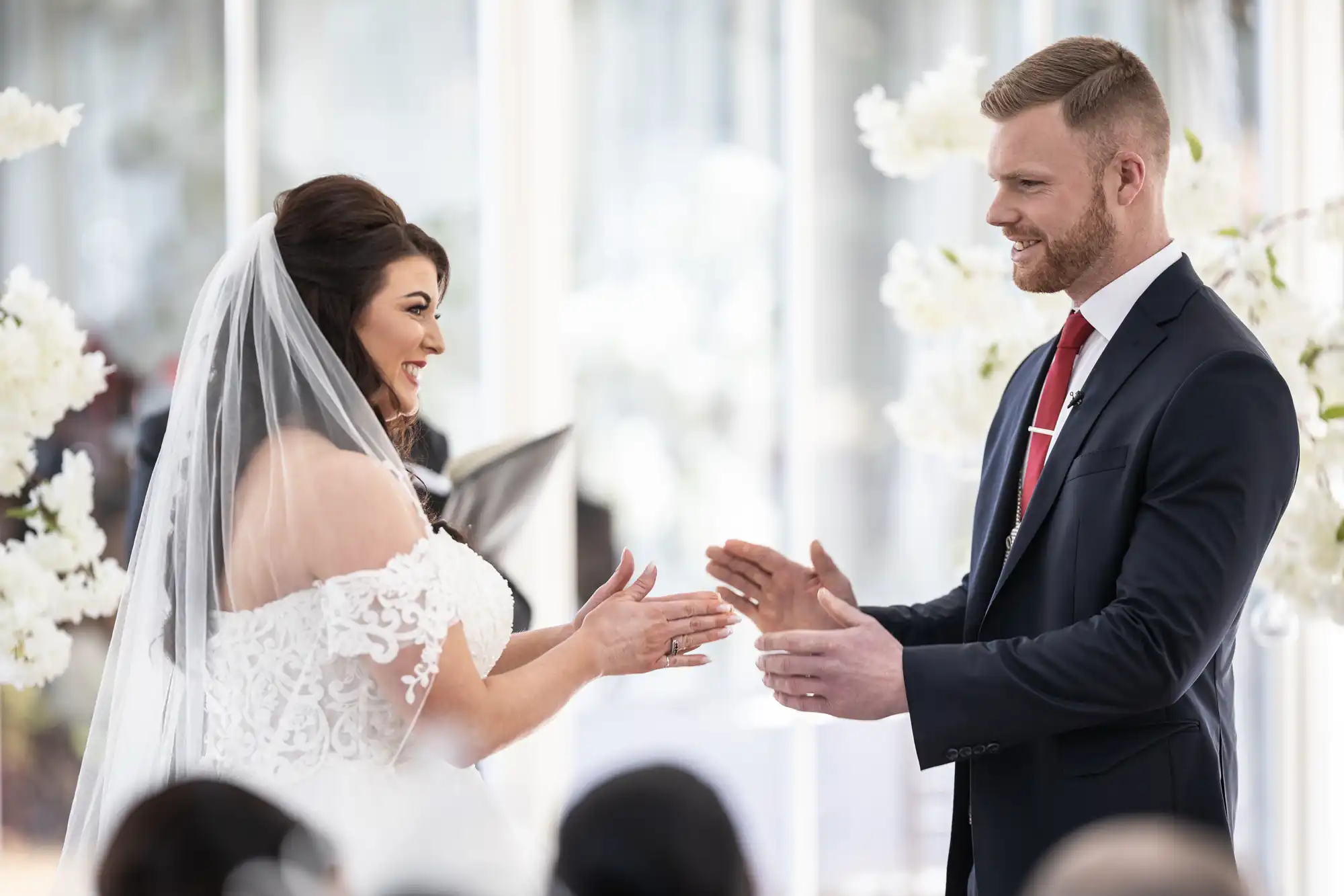 A bride and groom, both smiling, face each other at their wedding ceremony. The bride wears a white lace off-the-shoulder gown and a veil, while the groom is in a dark suit with a red tie.
