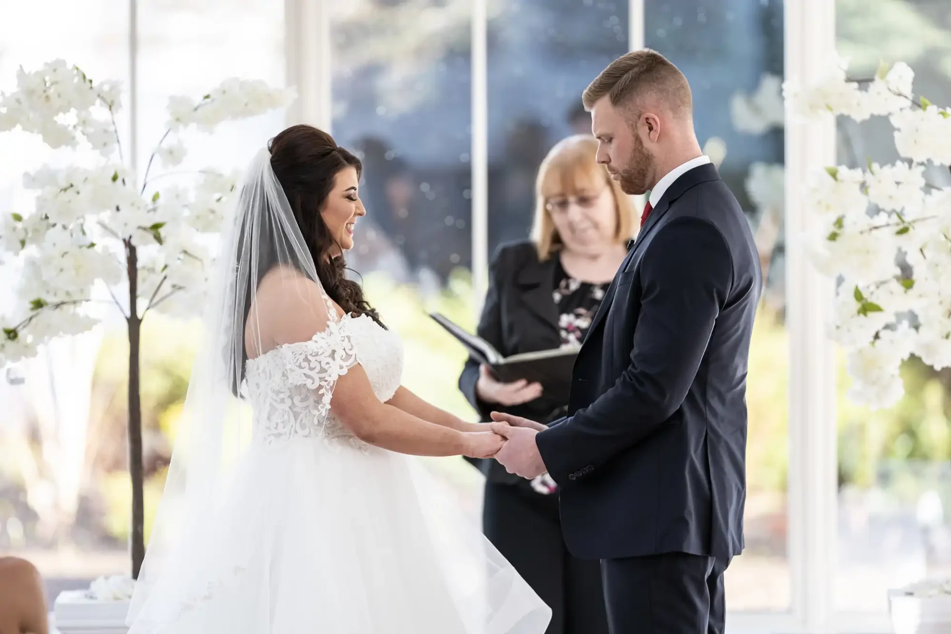 Bride and groom holding hands during a wedding ceremony in a bright room with white floral decorations, officiant in the background.