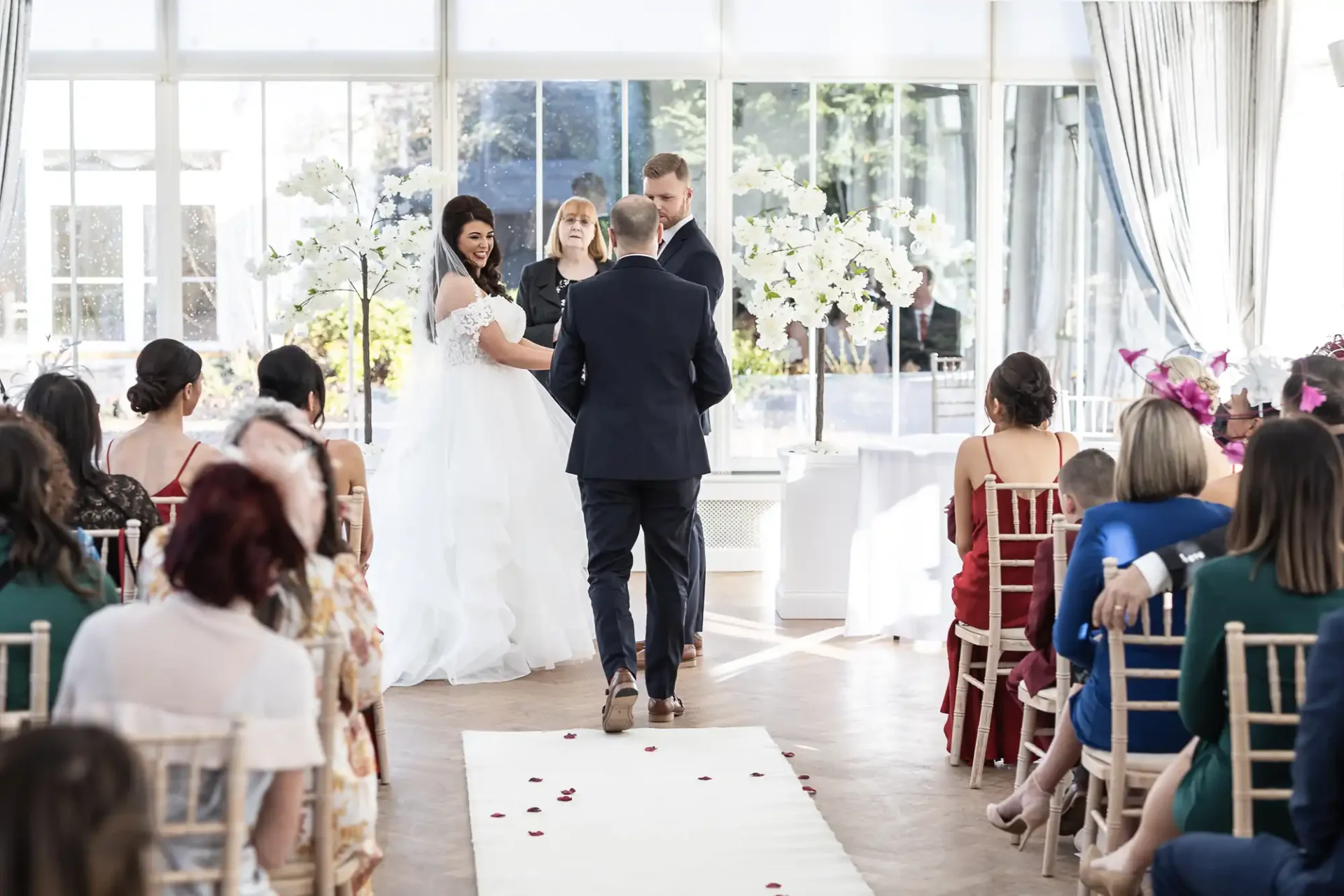 A bride and groom walking down the aisle, hand in hand, at their wedding ceremony in a bright, window-lined room filled with guests.