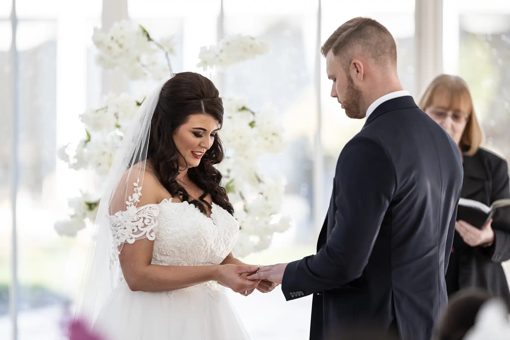 A bride placing a ring on the groom's finger during a wedding ceremony, with an officiant holding a book in the background.