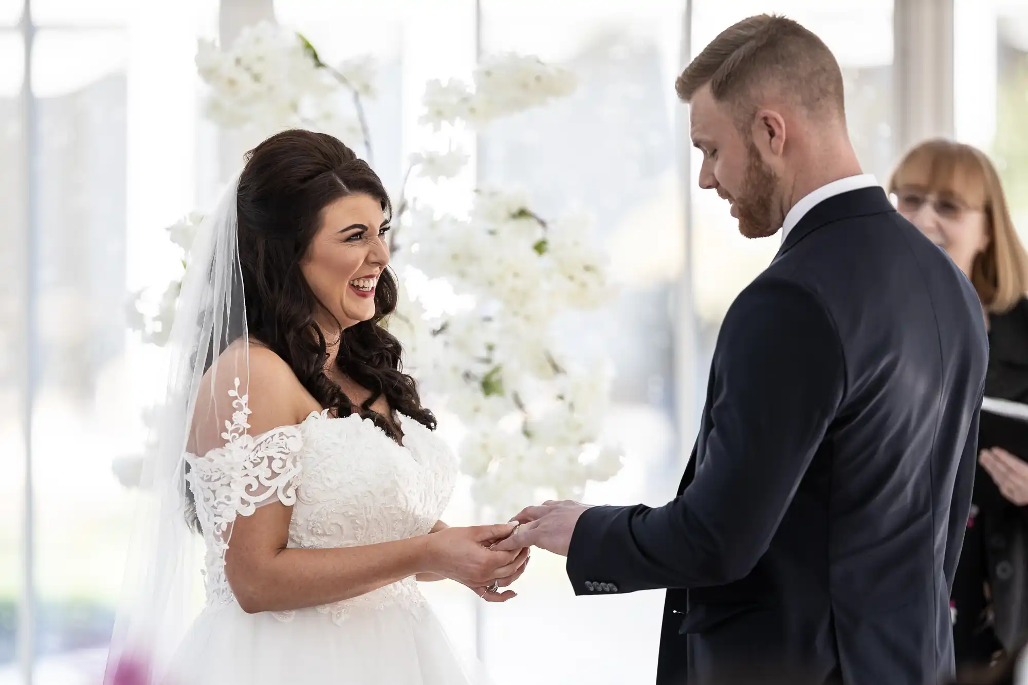 A bride and groom stand facing each other during a wedding ceremony, with the bride smiling as the groom places a ring on her finger. An officiant stands in the background holding a book.