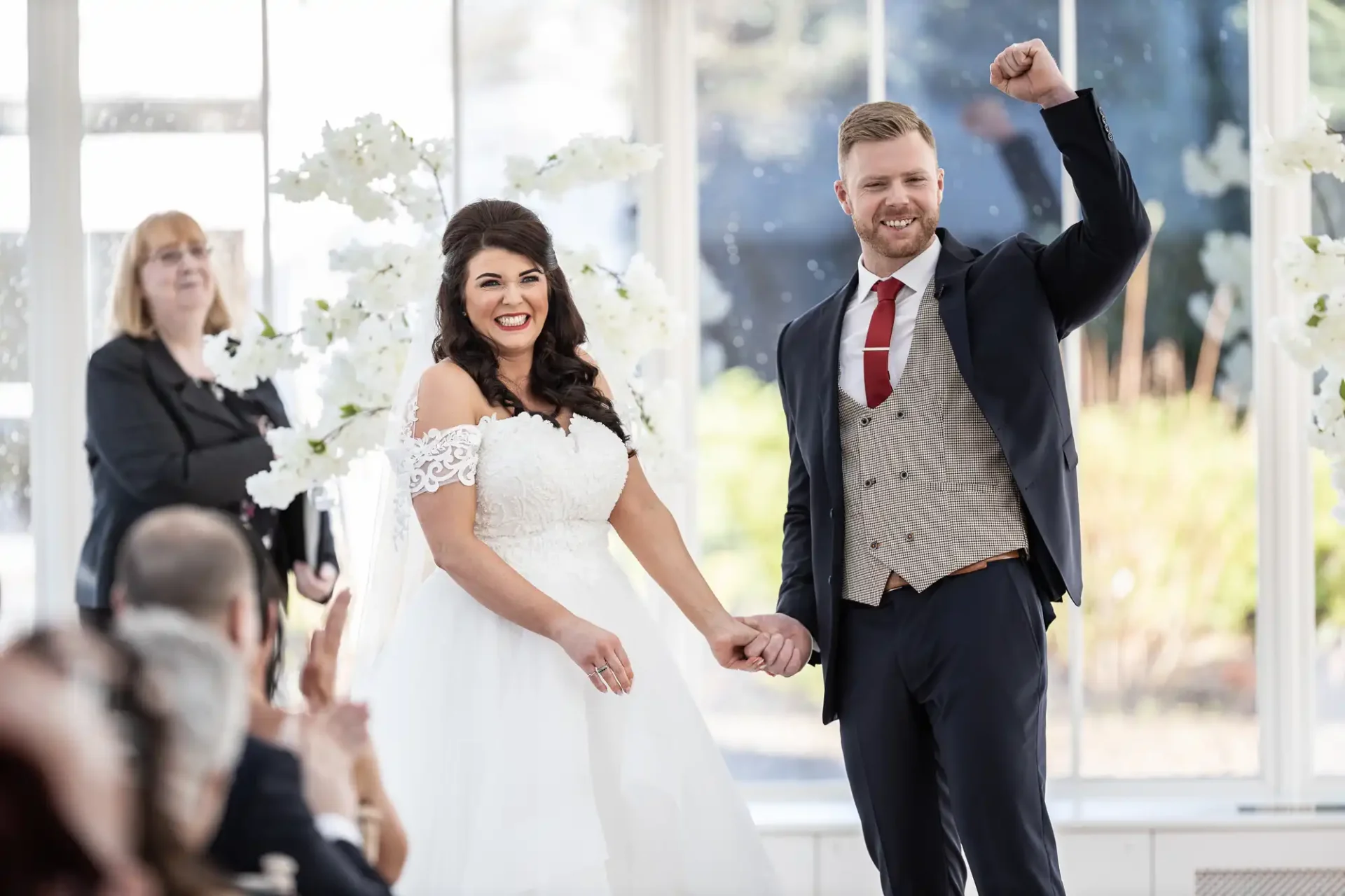 A bride and groom smiling and holding hands, the groom raising one fist in triumph, in a sunlit wedding venue with guests and an officiant in the background.