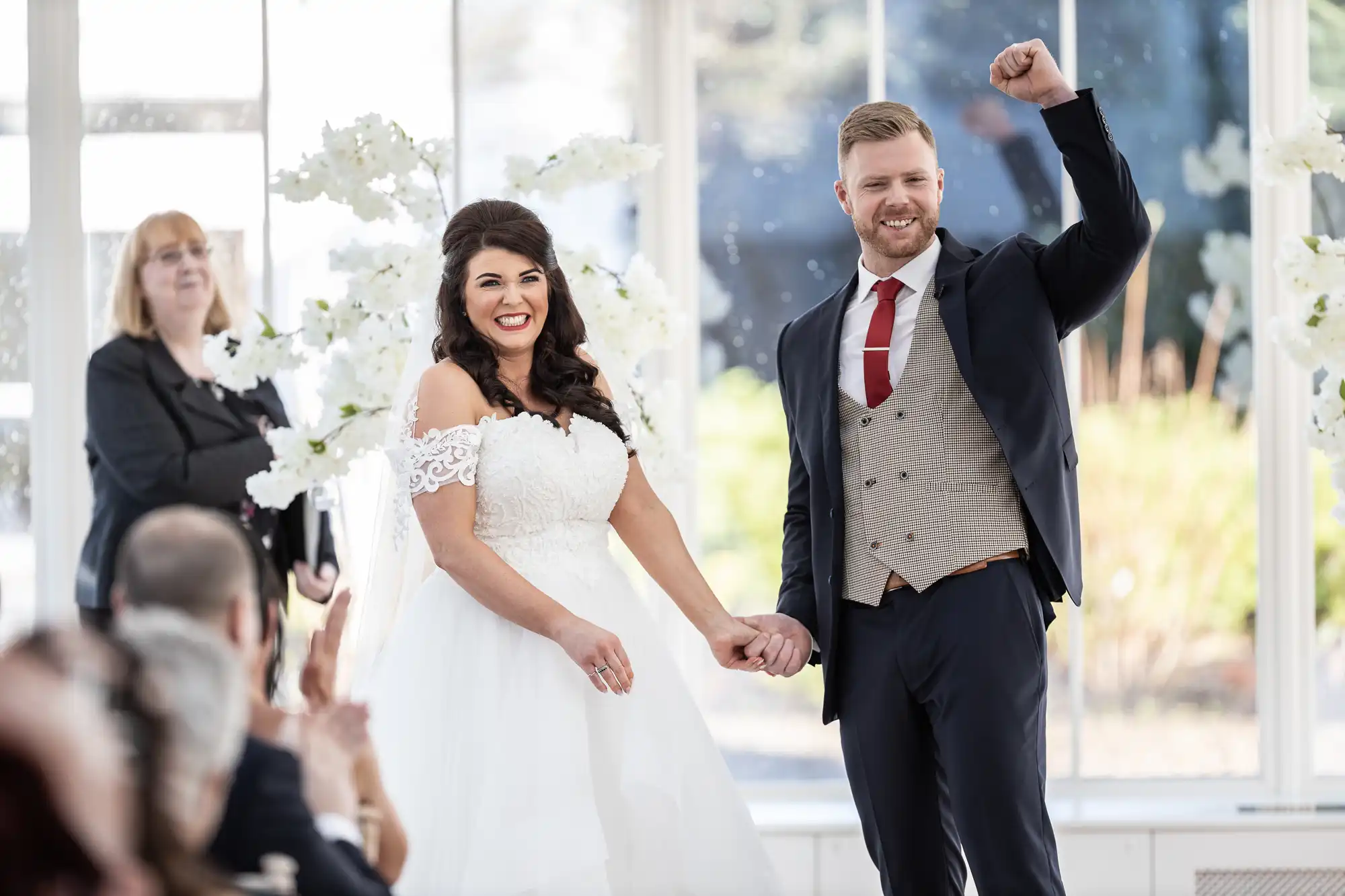 A bride and groom stand hand in hand, smiling, with the groom raising his fist in celebration. They are indoors with natural light and a backdrop of white flowers. Applauding guests are visible in the foreground.
