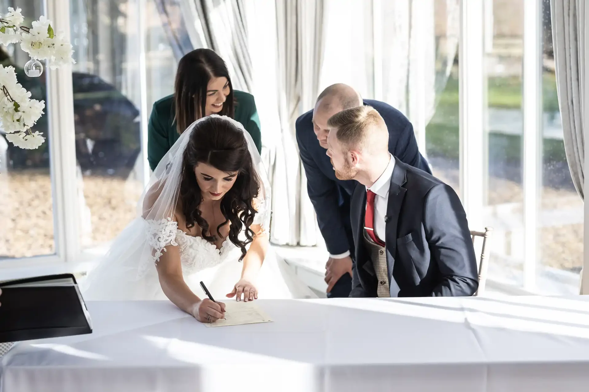 A bride in a white gown and a groom in a red tie and black suit watch as she signs a document at a table, with two witnesses standing behind them.