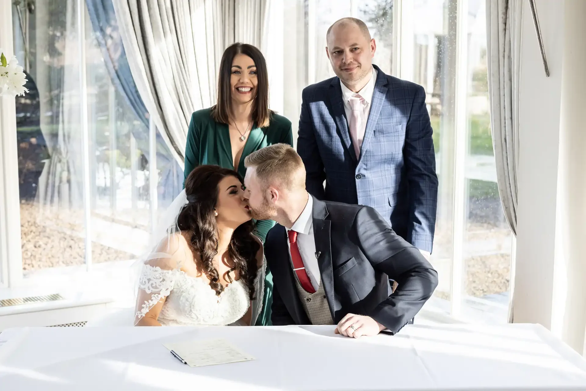 A bride in a white dress kisses a groom in a suit at a signing table during a wedding ceremony, with a smiling officiant standing behind them.