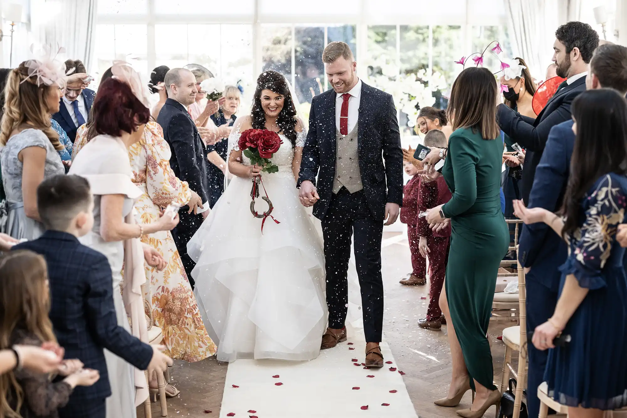 A bride and groom walk down the aisle holding hands, surrounded by guests celebrating and taking photos. The bride holds a bouquet of red roses, and flower petals are scattered on the floor.